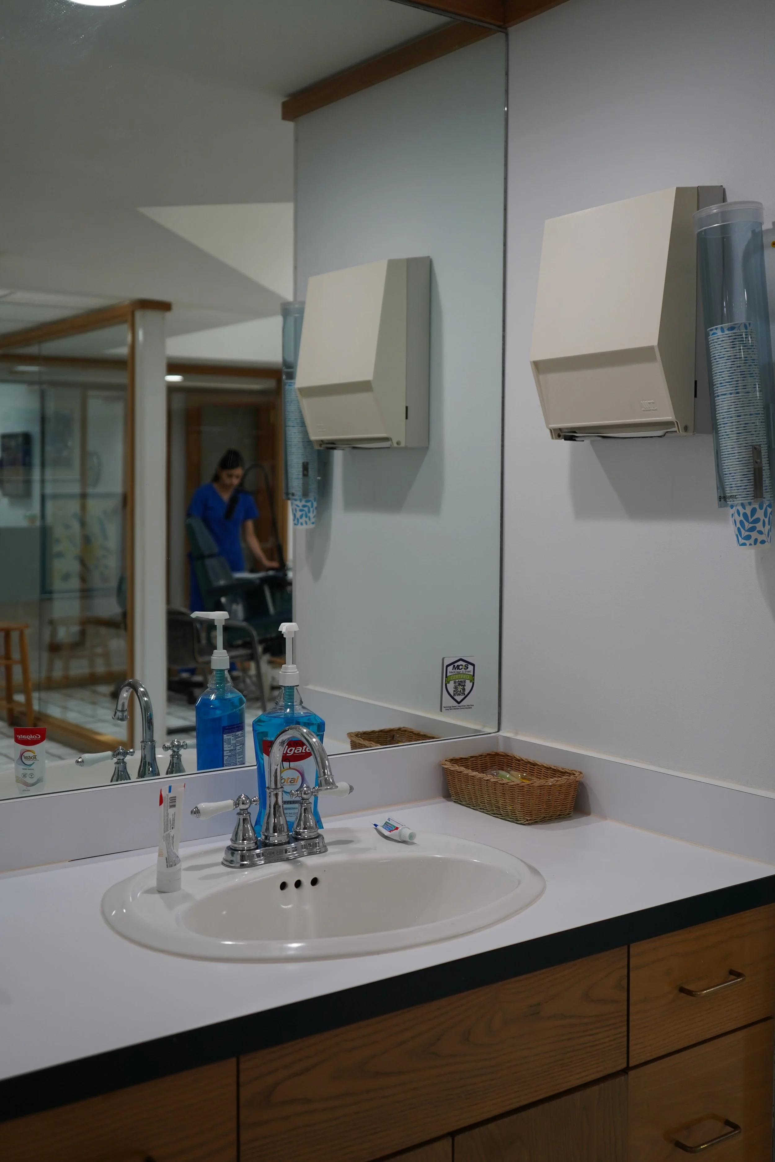 Bathroom sink with soap and toothbrushes, large mirror, paper towel dispenser, and wall-mounted hand sanitizer bottle, with a nurse in blue scrubs visible in the background.