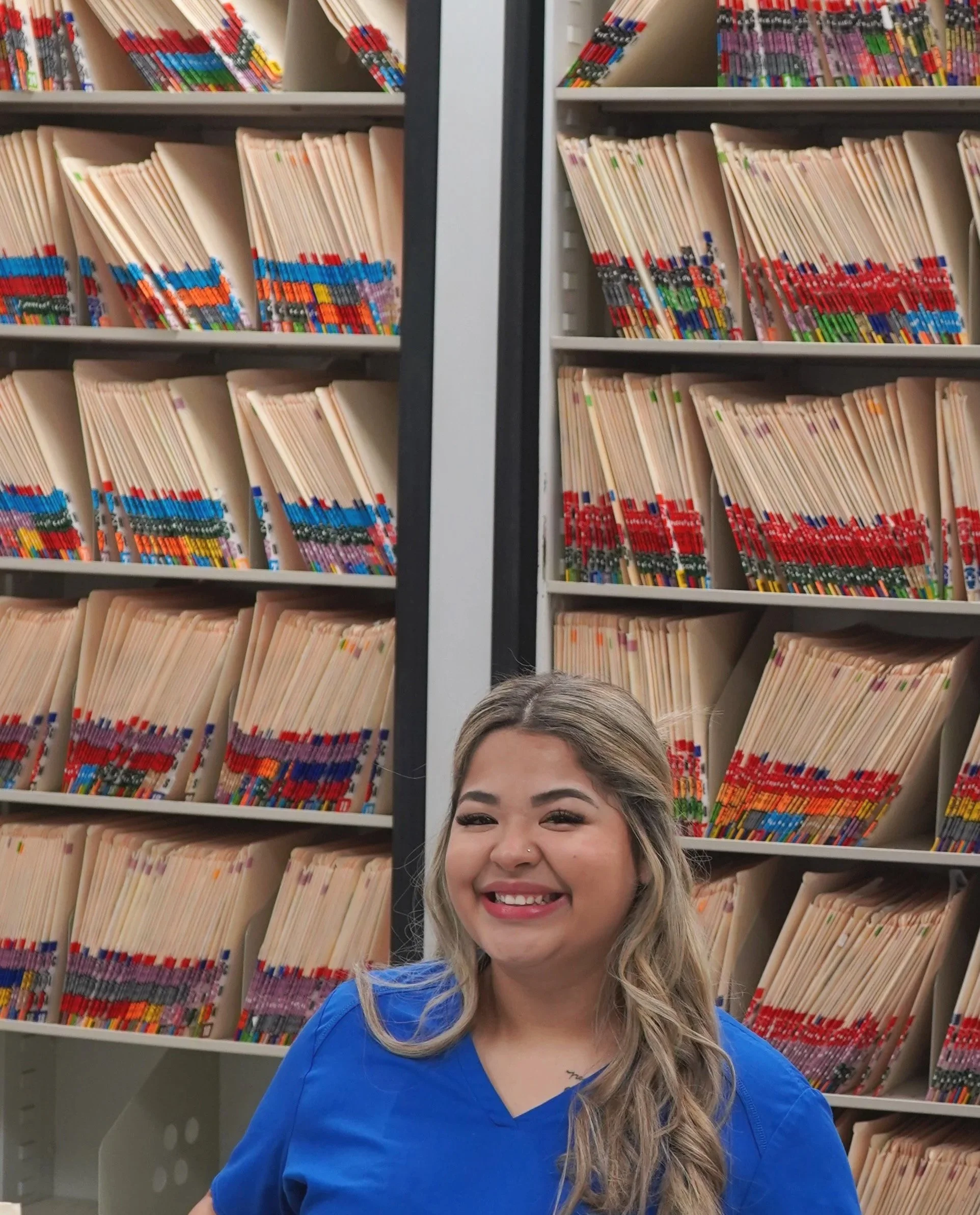 A smiling woman with long blonde hair wearing a blue shirt standing in front of shelves filled with beige filing folders marked with colored labels in a library or office.