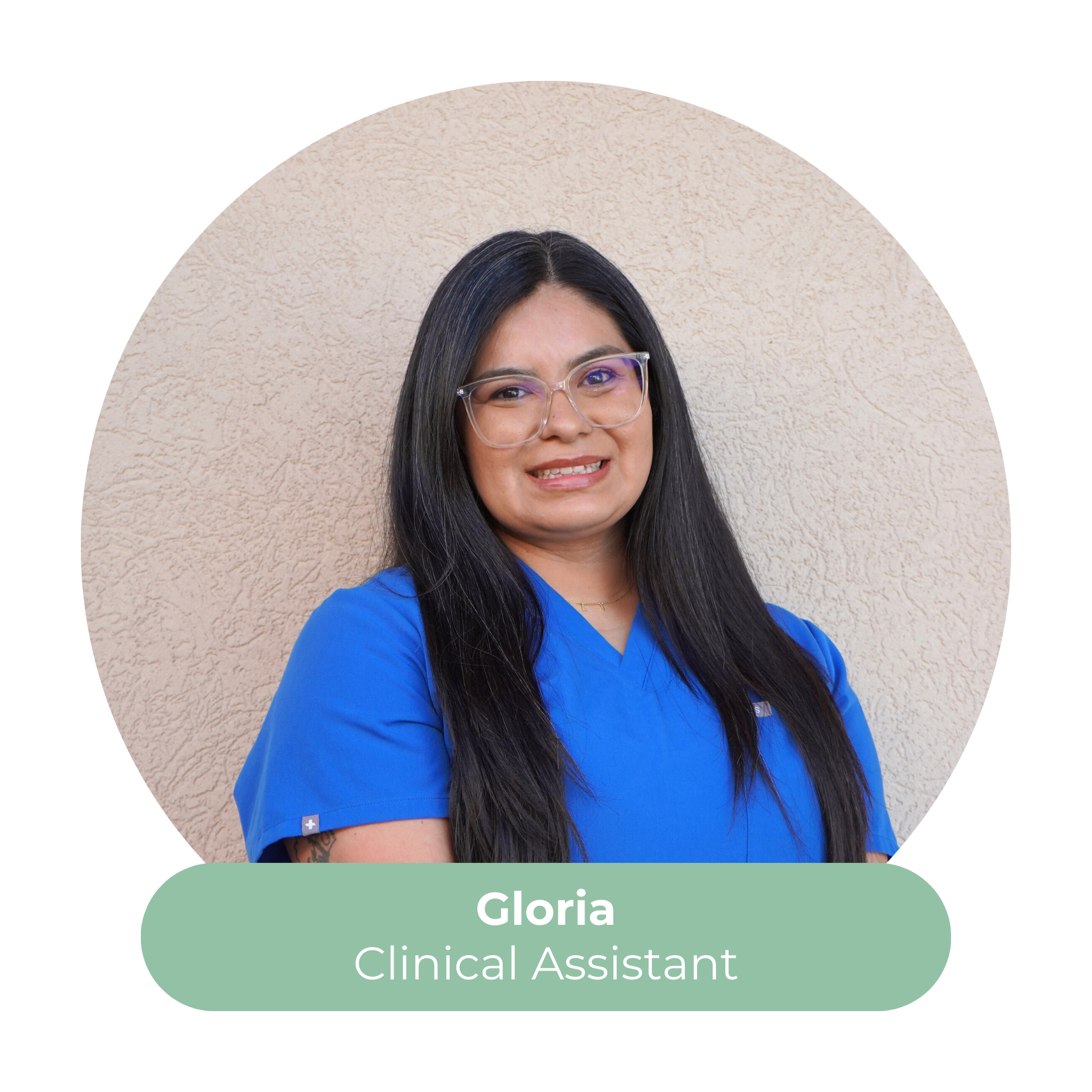 Portrait of Gloria, a smiling woman with long black hair and glasses, wearing blue medical scrubs, standing against a beige wall. Caption reads: Gloria, Clinical Assistant.