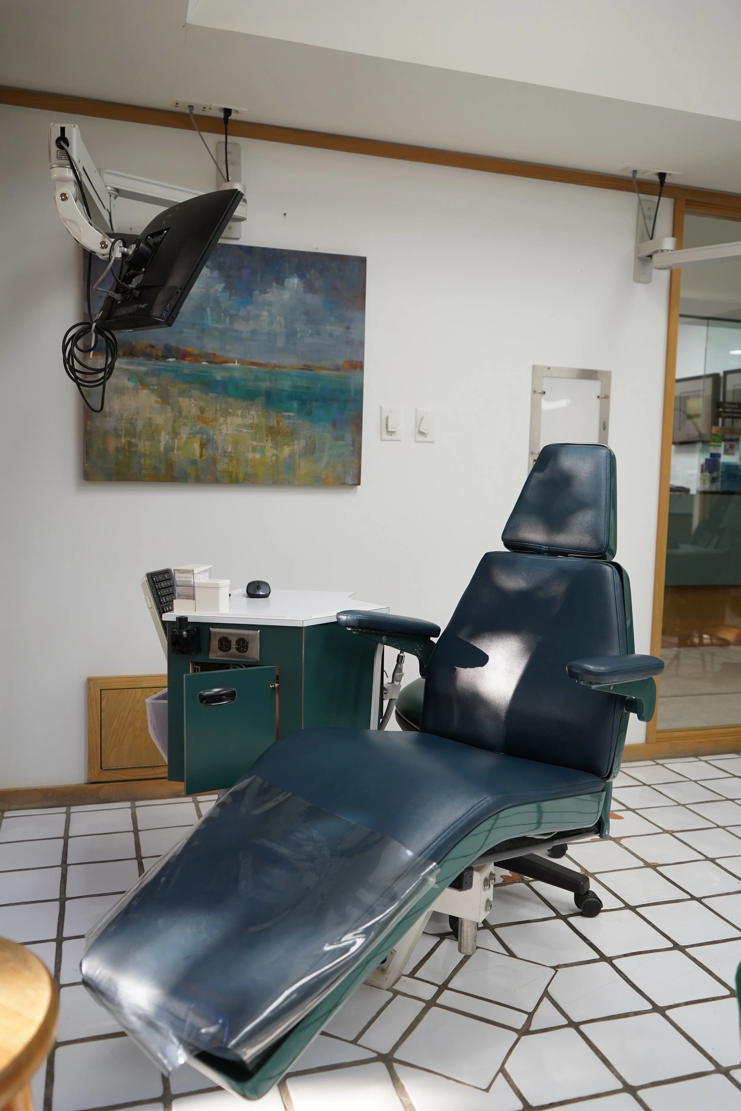Medical examination room with a reclining chair, a small side table, a wall-mounted monitor, and a painting of a landscape.
