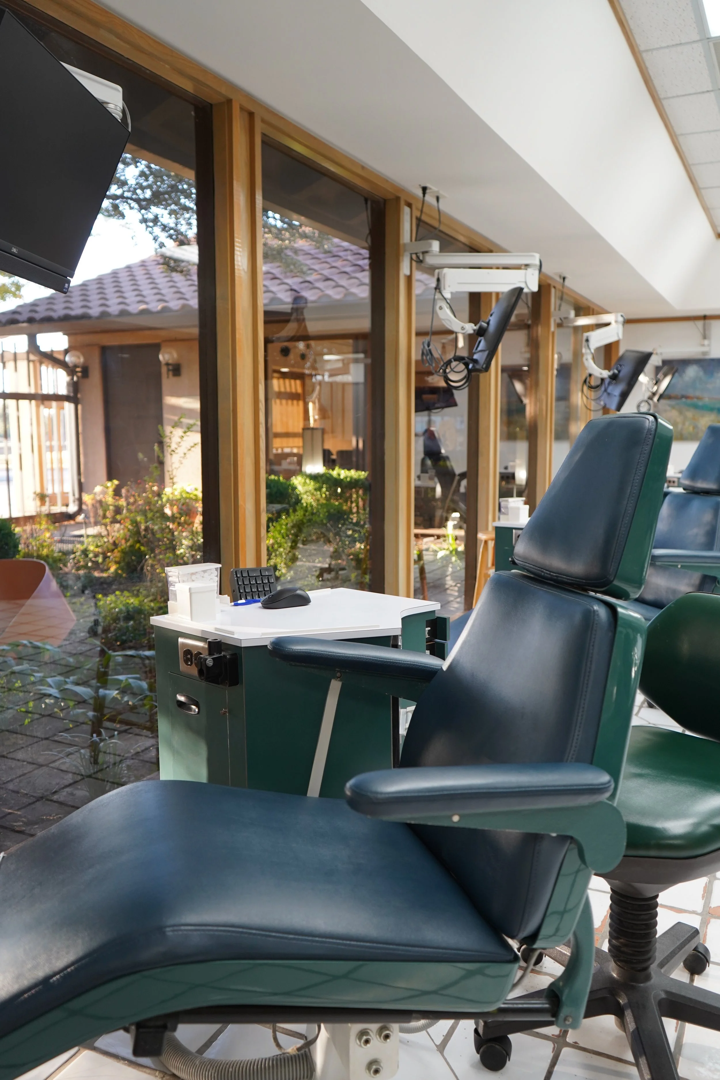 Empty medical examination chair in a clinical setting near large windows and some medical equipment.
