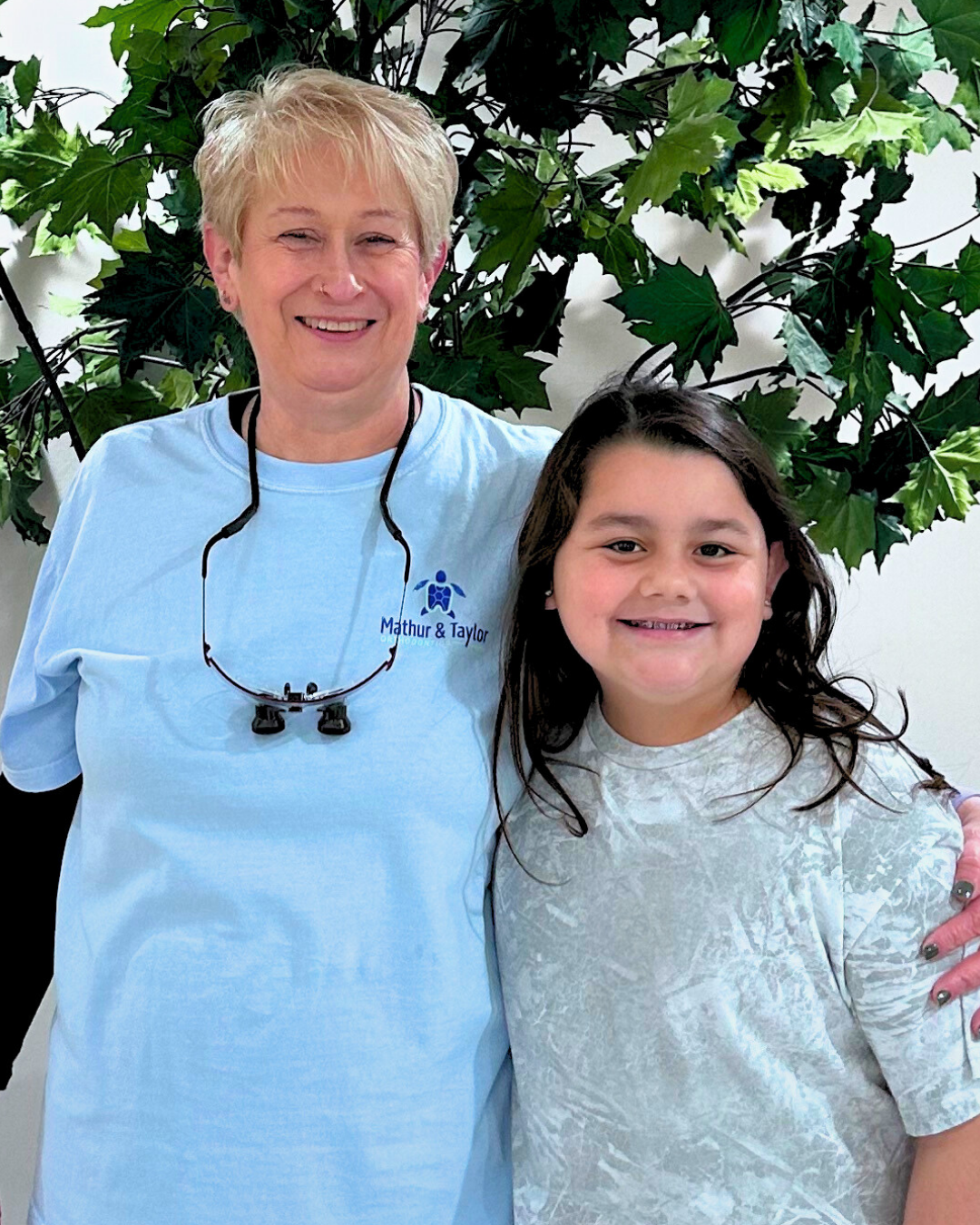 Two smiling females, one adult and one child, standing in front of a leafy green backdrop, with the adult wearing a light blue shirt with a logo that reads 'Mathur & Taylor'.