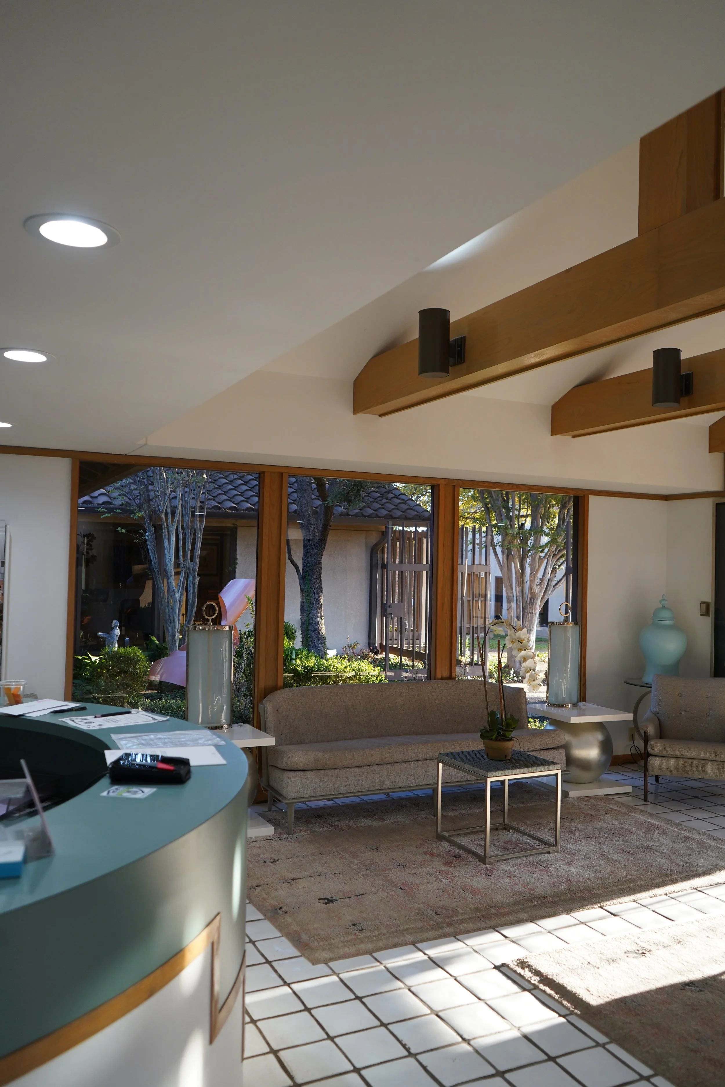 Interior view of a modern waiting area with beige sofas, a small coffee table with a potted plant, large windows revealing a garden, and decorative vases.
