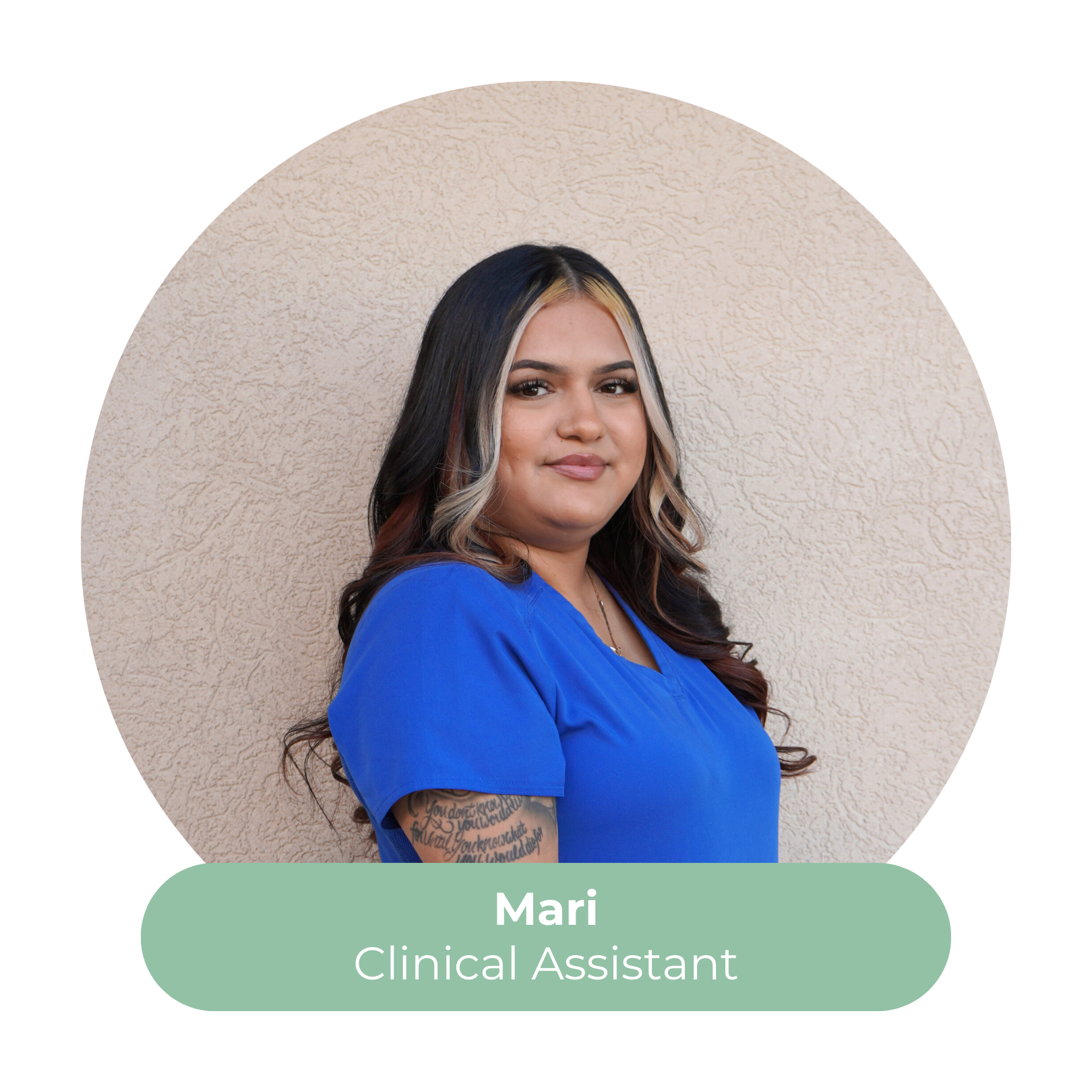 Portrait of Mari, a clinical assistant, wearing a blue uniform, with long dark hair and a tattoo on her left arm, standing against a beige textured wall.