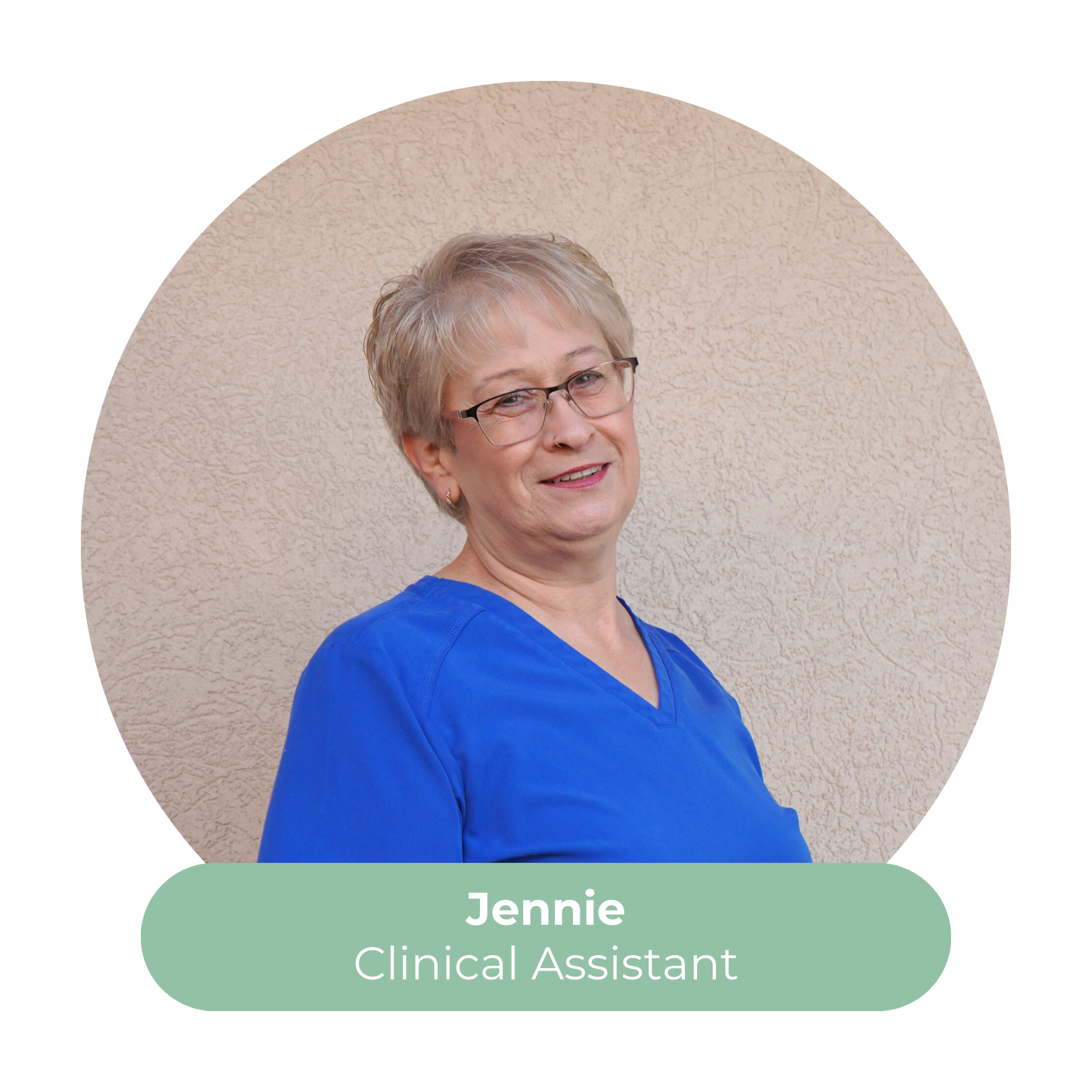 Portrait of Jennie, a smiling middle-aged woman with short gray hair, wearing glasses and blue scrubs, standing against a beige wall, professional appearance, identified as a clinical assistant.