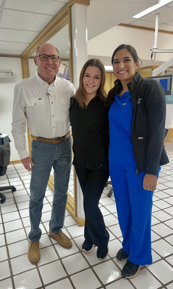 Three people standing together indoors, smiling at the camera. The man on the left is wearing glasses, a white button-up shirt, and jeans. The woman in the middle is wearing a black jacket and dark pants. The woman on the right is dressed in blue scrubs and a black jacket, indicating she might be a healthcare worker.
