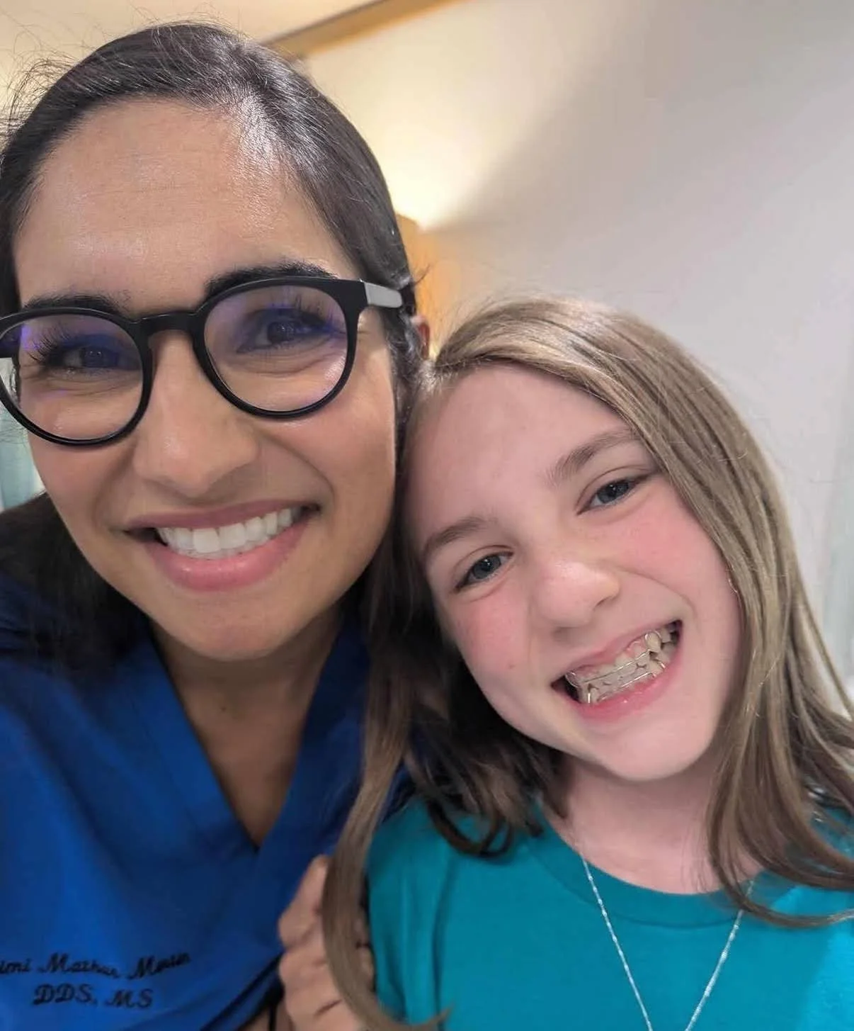 A woman with glasses and a young girl smiling together, both showing their teeth, in an indoor setting.