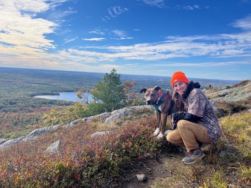 Photo of Sam kneeling next to same dog at a hike viewpoint overlooking a lake