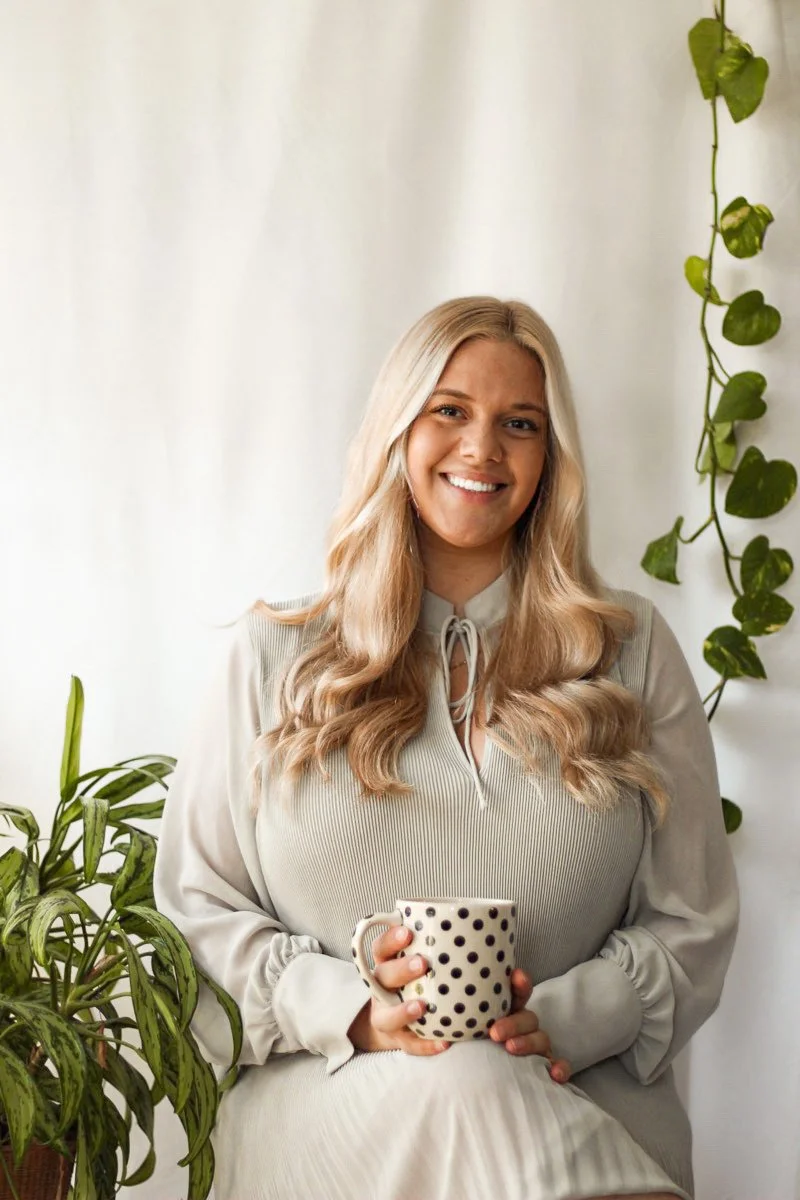 Une femme blonde souriante, assise près d'une plante, tenant une tasse à pois noirs.