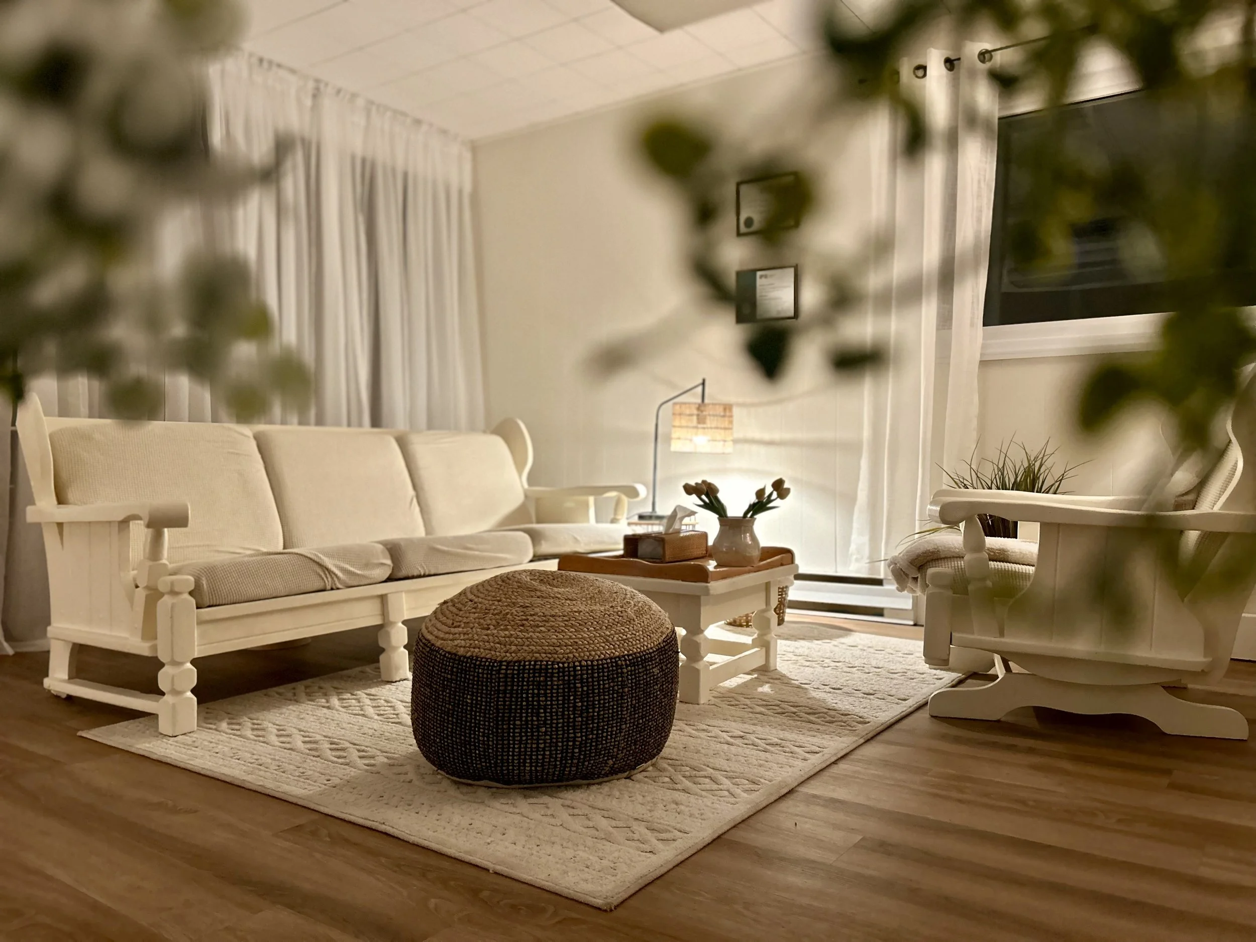 Salon chaleureux avec canapé et fauteuil blancs, tapis beige, pouf rond marron clair, table basse en bois avec fleurs, lampadaire et télévision, fenêtres avec rideaux blancs, décoration sobre et cosy.