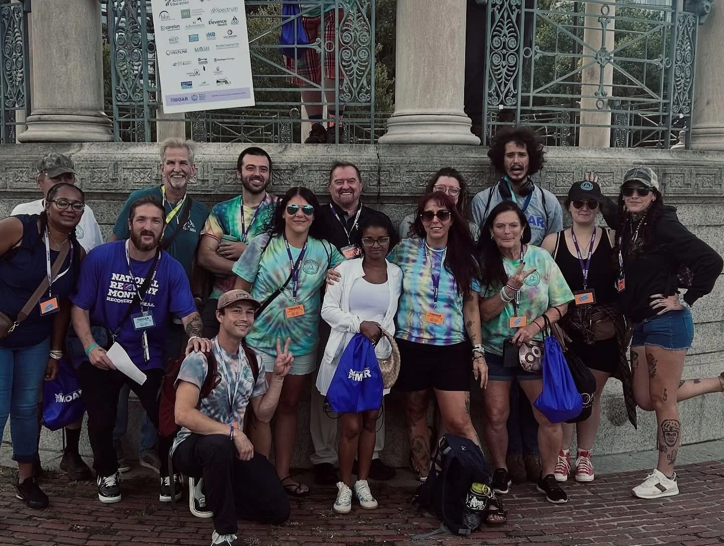 Group of people posing outdoors in front of a stone and iron structure, some are wearing tie-dye shirts and ID badges.