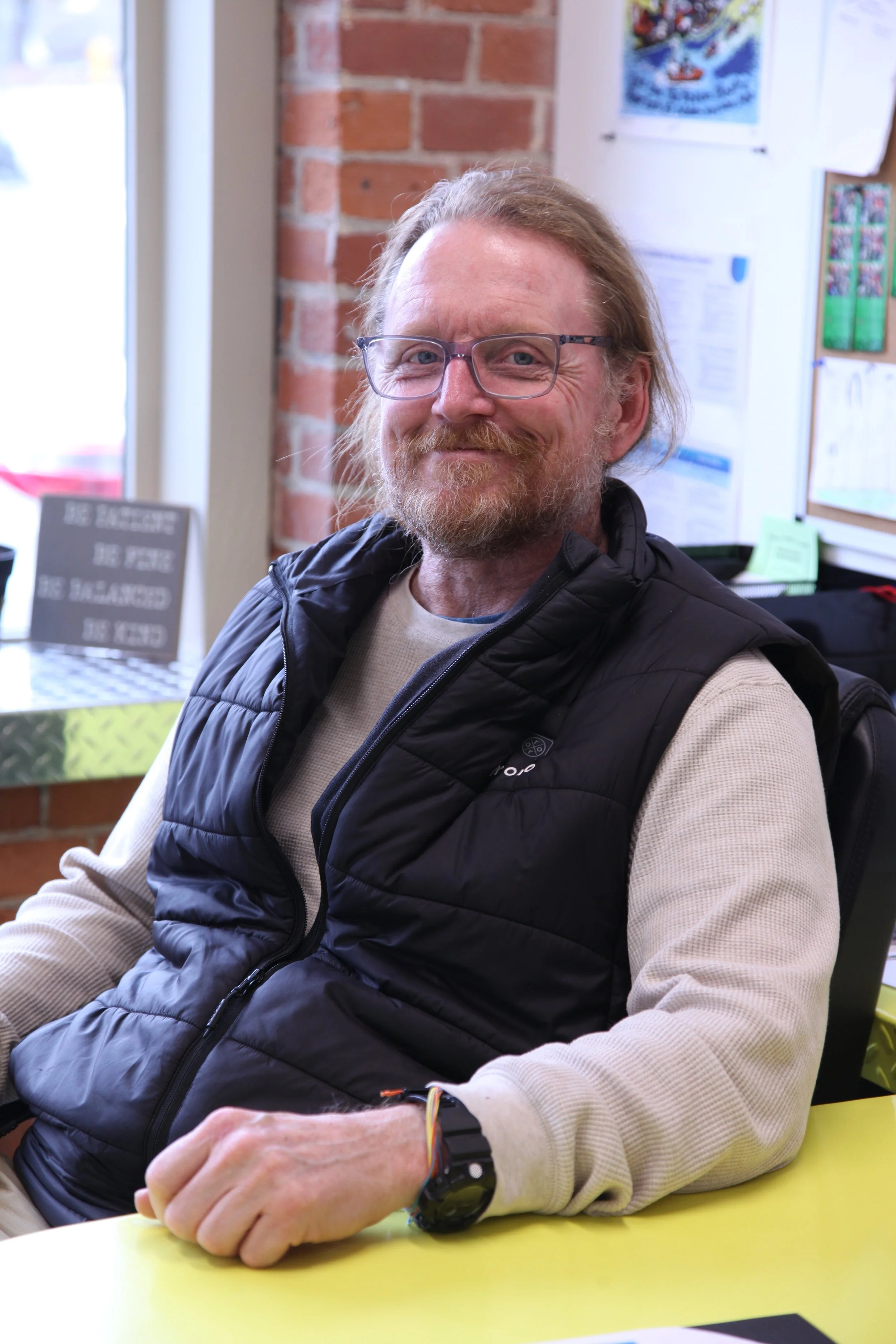 A man with glasses and a beard, smiling and sitting at a yellow table inside a room with a brick wall and various posters and papers in the background.