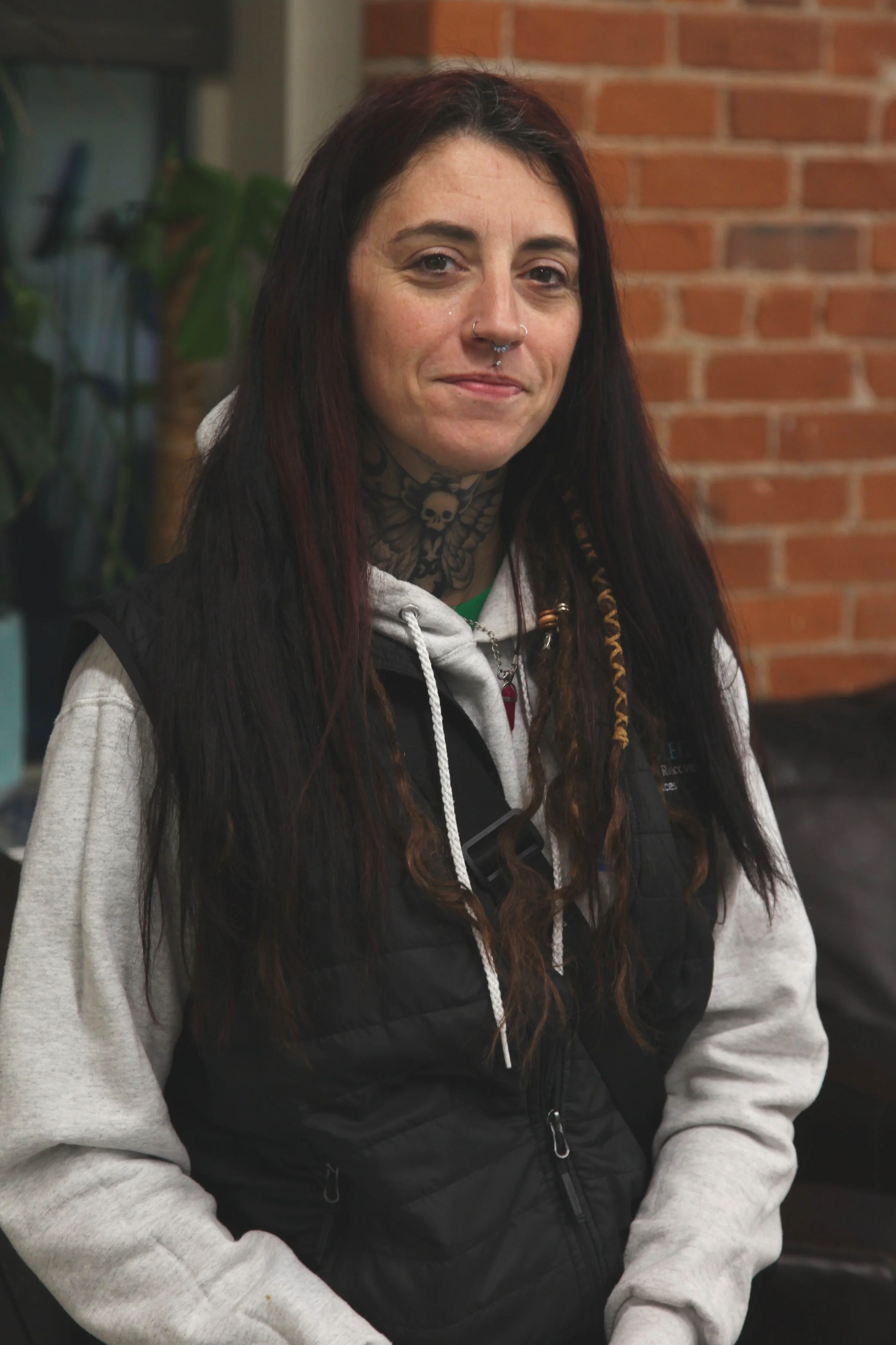 Young woman with dark hair and tattoos, wearing a hoodie and vest, standing indoors with a brick wall in the background.