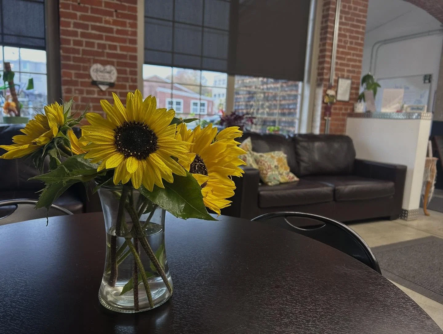 A bouquet of bright yellow sunflowers in a clear glass vase on a dark wooden table inside a room with brick walls and large window panes.