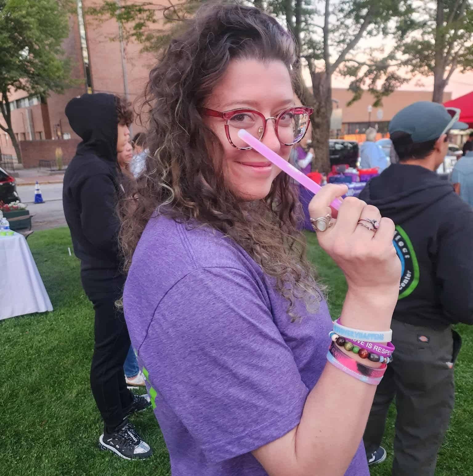 A woman with curly hair wearing glasses, a purple shirt, and bracelets, smiling and holding a pink glow stick, at an outdoor gathering with other people and tables in the background.