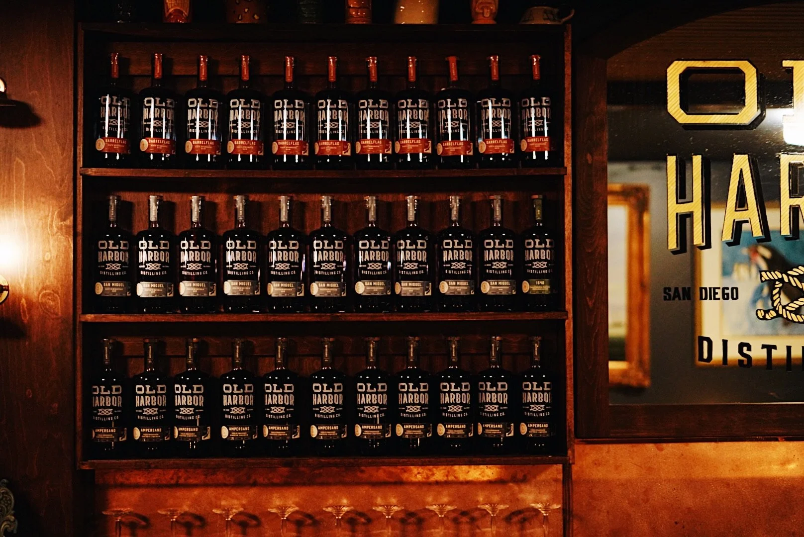 Shelf with multiple bottles of Old Harbor Distilling Co. liquor brands, arranged by label color, in a bar or store setting.