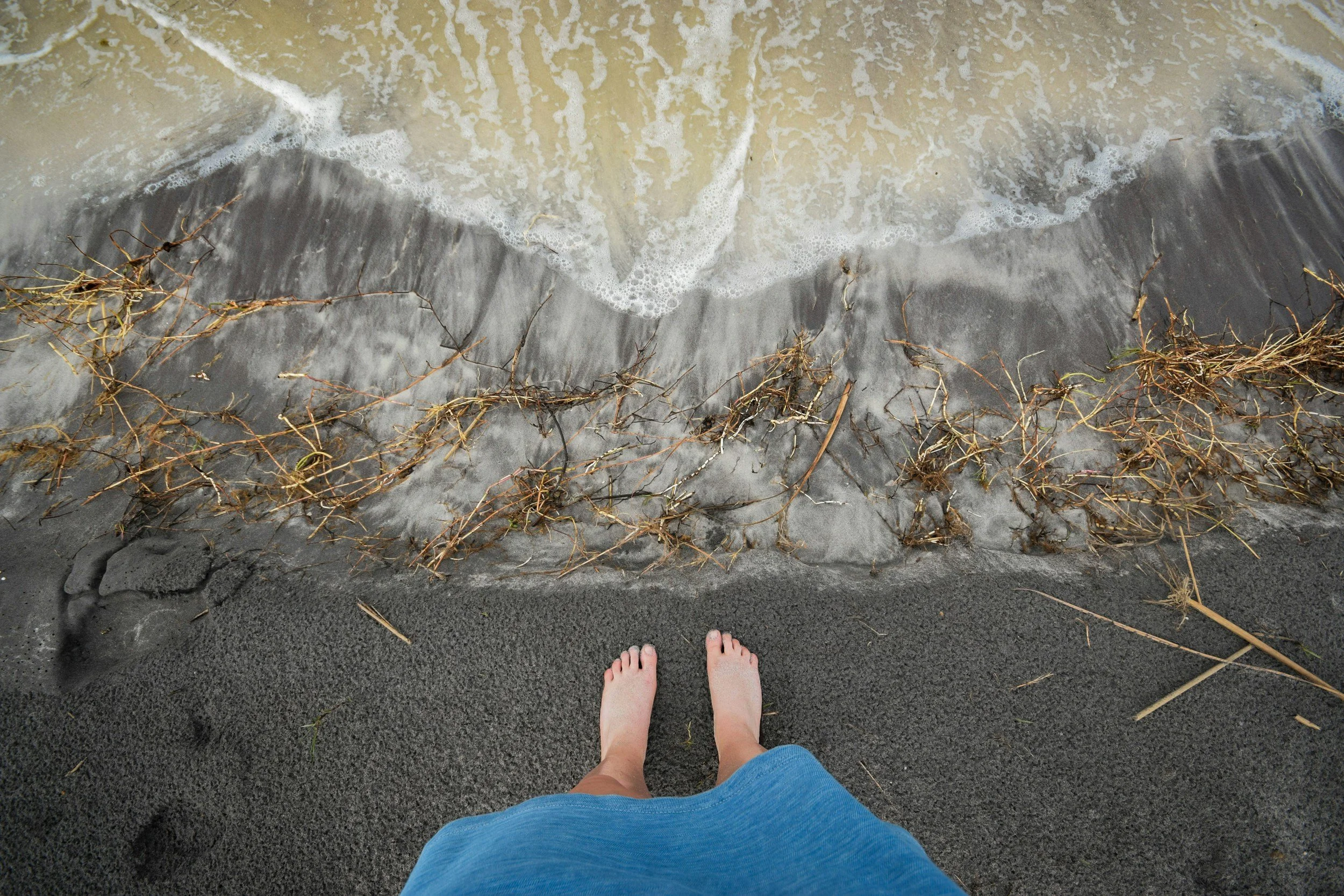 A person's feet standing on dark sand at the edge of a body of water with small waves touching the shore, some dried seaweed scattered on the sand.