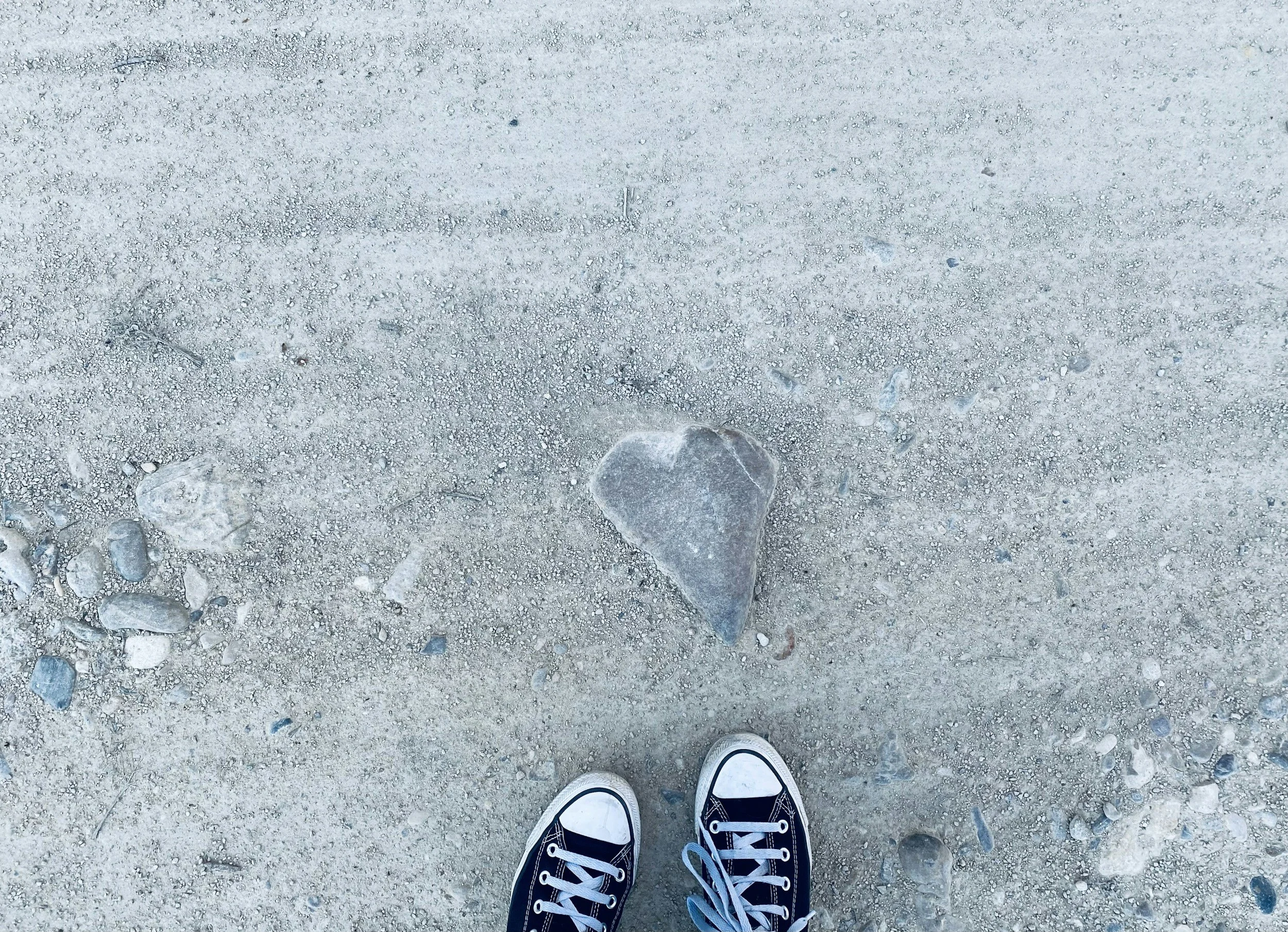 A pair of black sneakers with white laces and soles, standing on sandy ground with small rocks, and a heart-shaped stone on the sand.