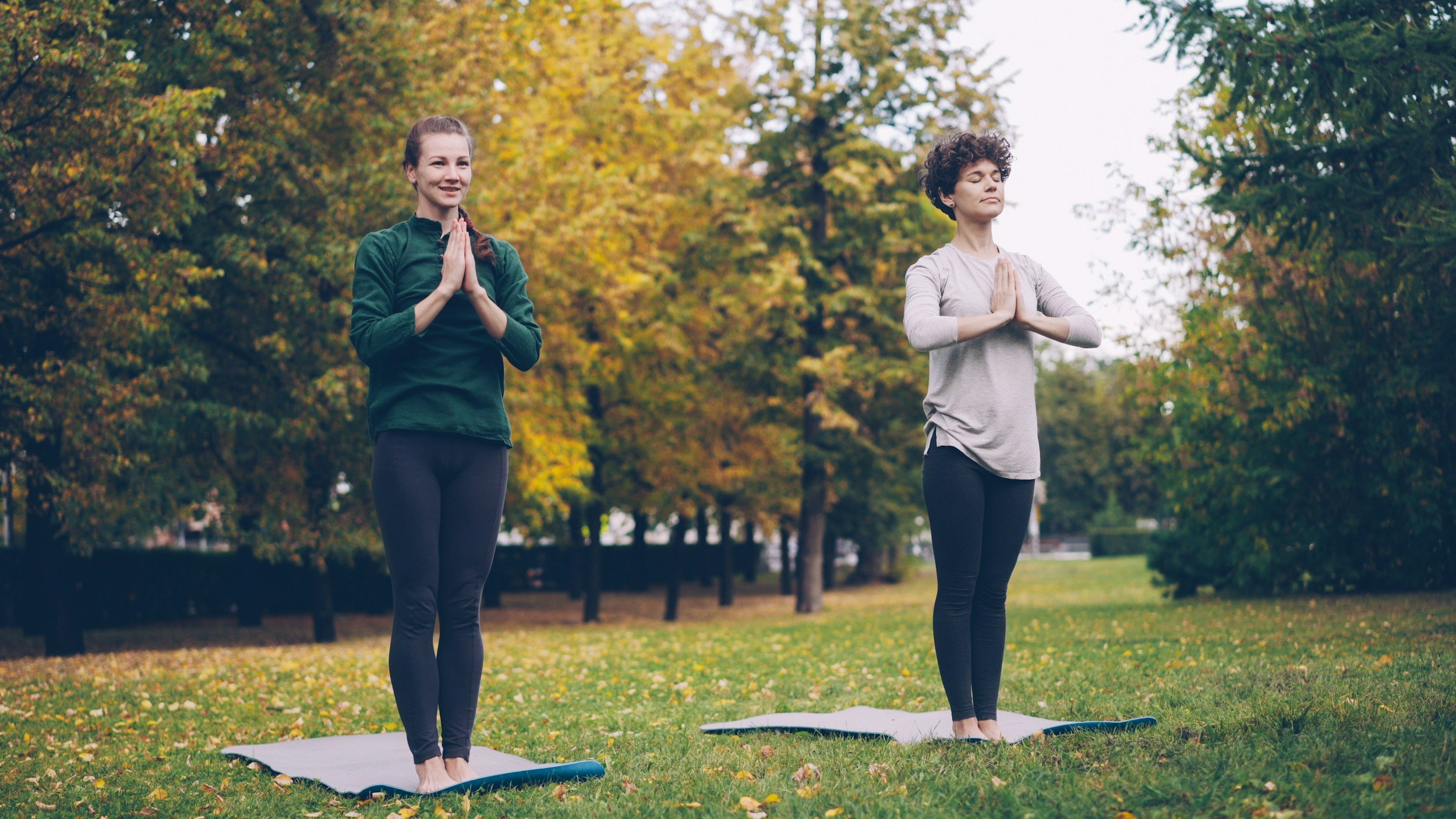 Two women practicing yoga outdoors on mats, standing with hands in prayer position, surrounded by autumn trees.