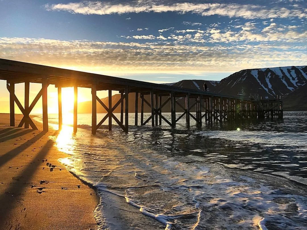 Sunset over a wooden pier extending into a body of water with mountains in the background.