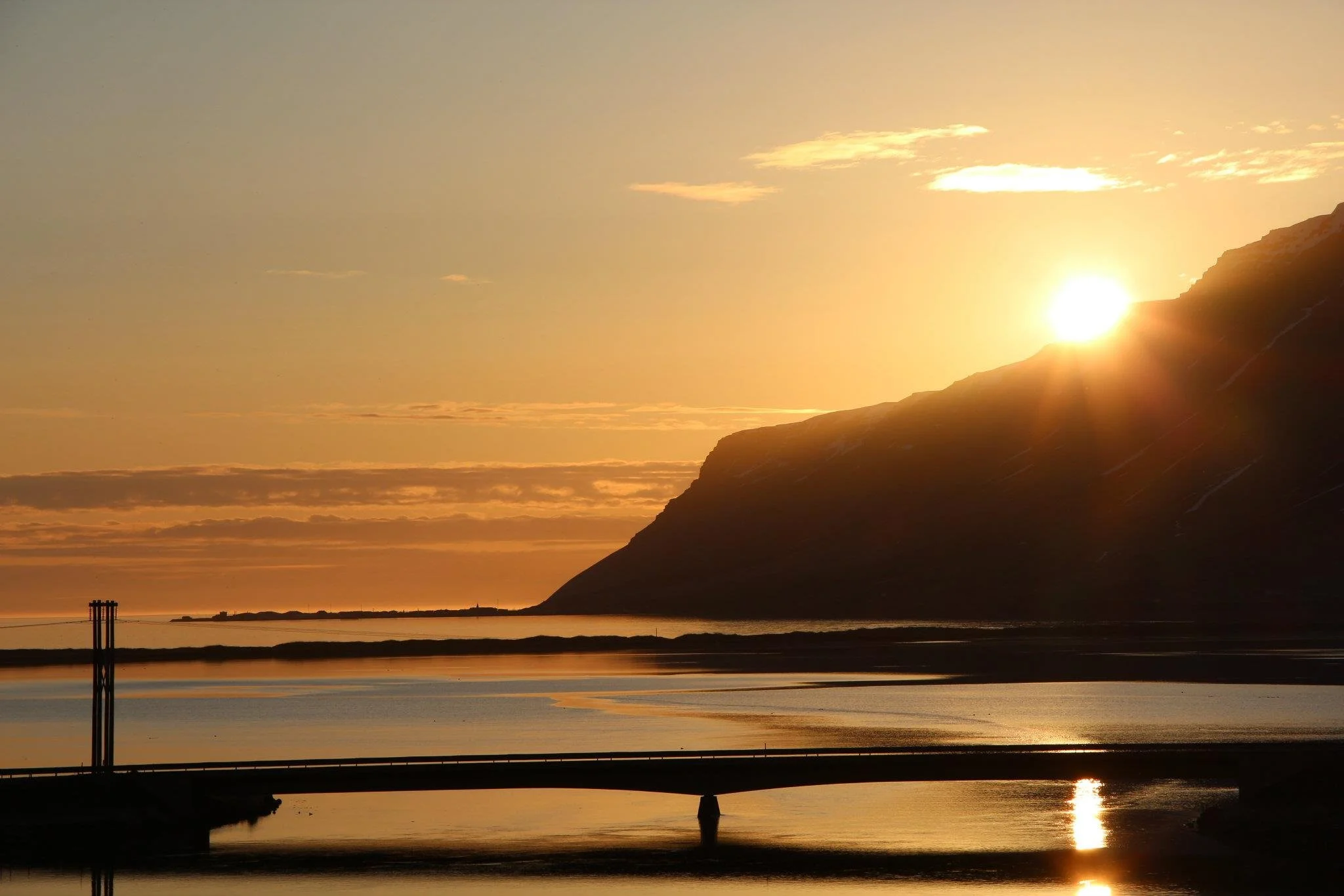 Sunset over a mountain and body of water, with a bridge in the foreground and a few clouds in the sky.