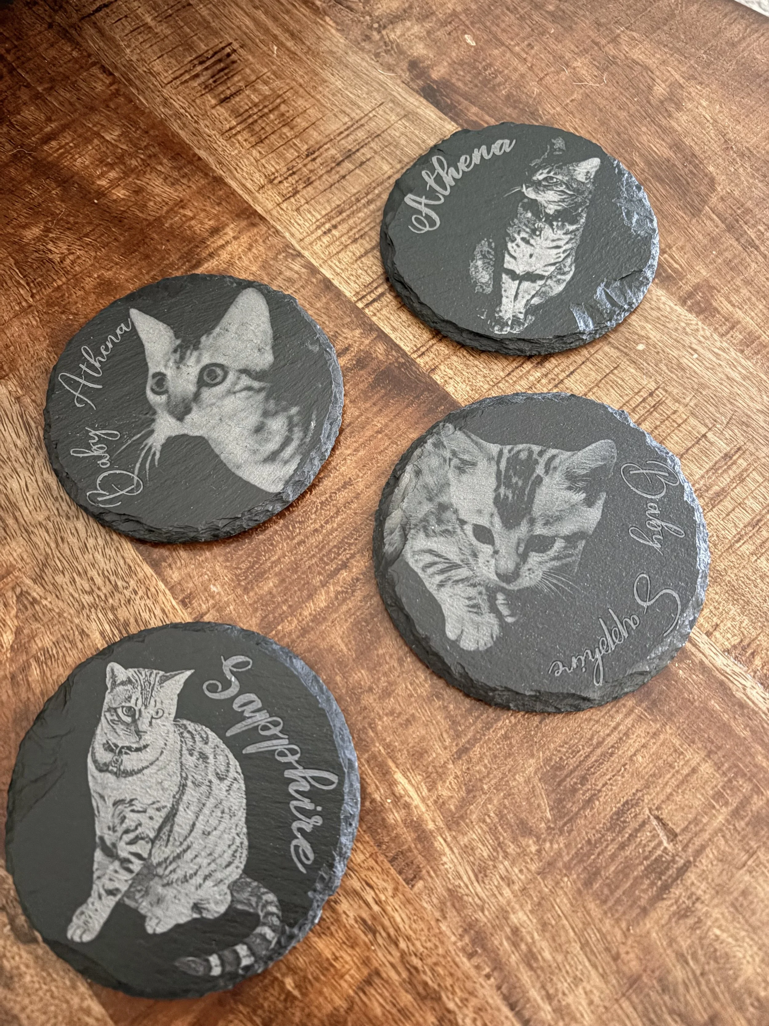 Four black slate coasters with engraved images of cats and the names 'Athena,' 'Athena,' 'Angelina,' and 'Sageblade,' placed on a wooden surface.