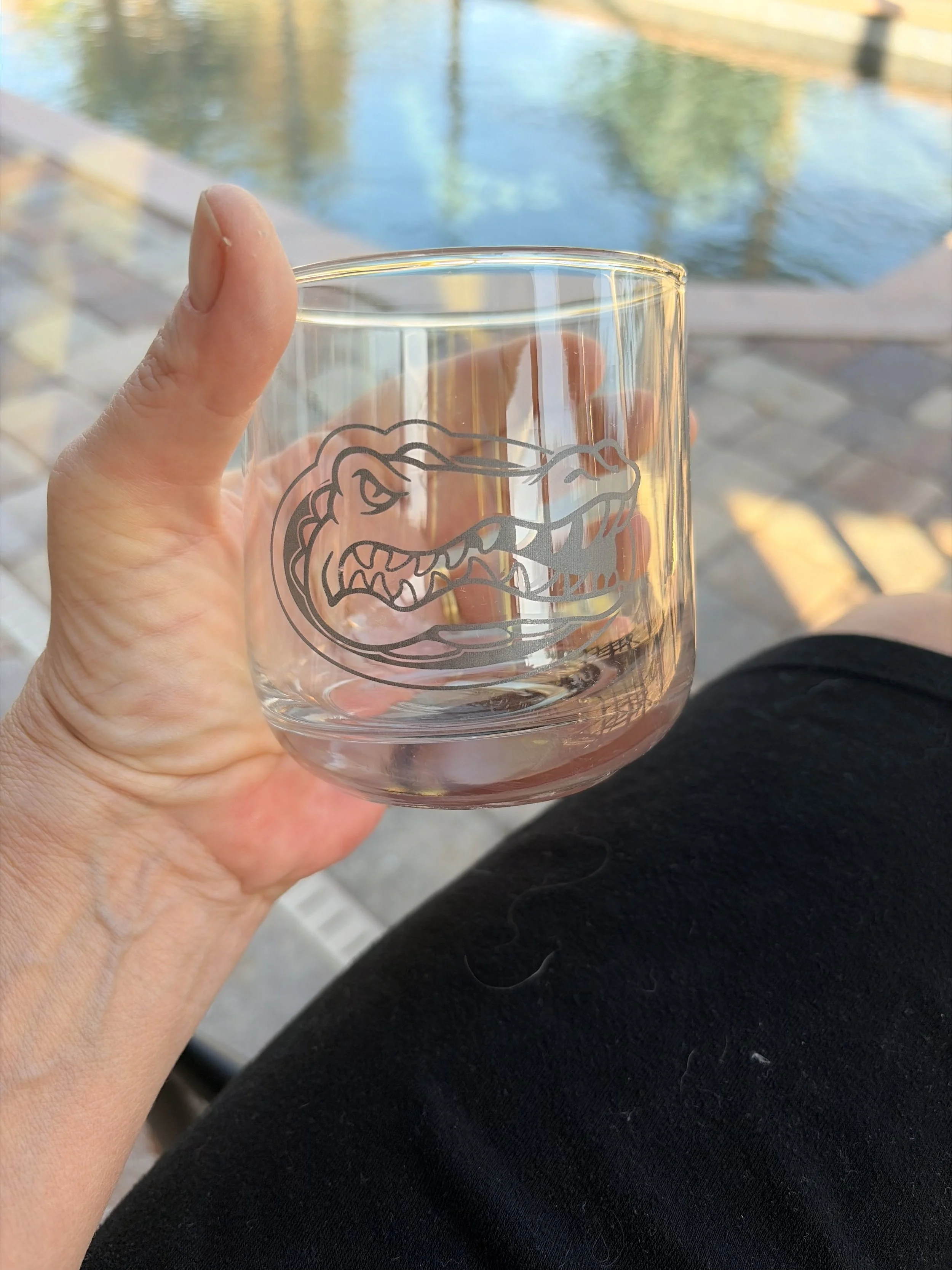 A clear glass with a crocodile logo held by a person's hand, with a water feature and brick patio in the background.