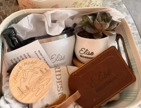 A tray holds two cups with succulents, a brown leather wallet, a small round wooden coaster, a white paper bag, a piece of paper, and a white paper bag with printed text.