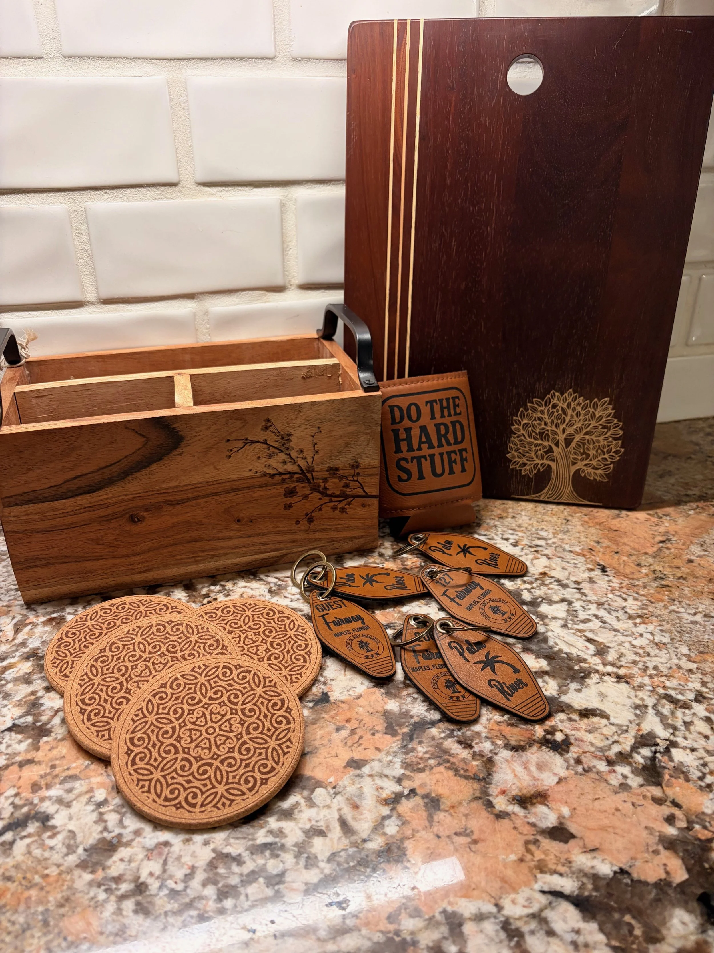 Wooden box with floral design, a sign that says 'Do the Hard Stuff,' a wooden board with tree engraving, and several keychains and coasters on a granite countertop.