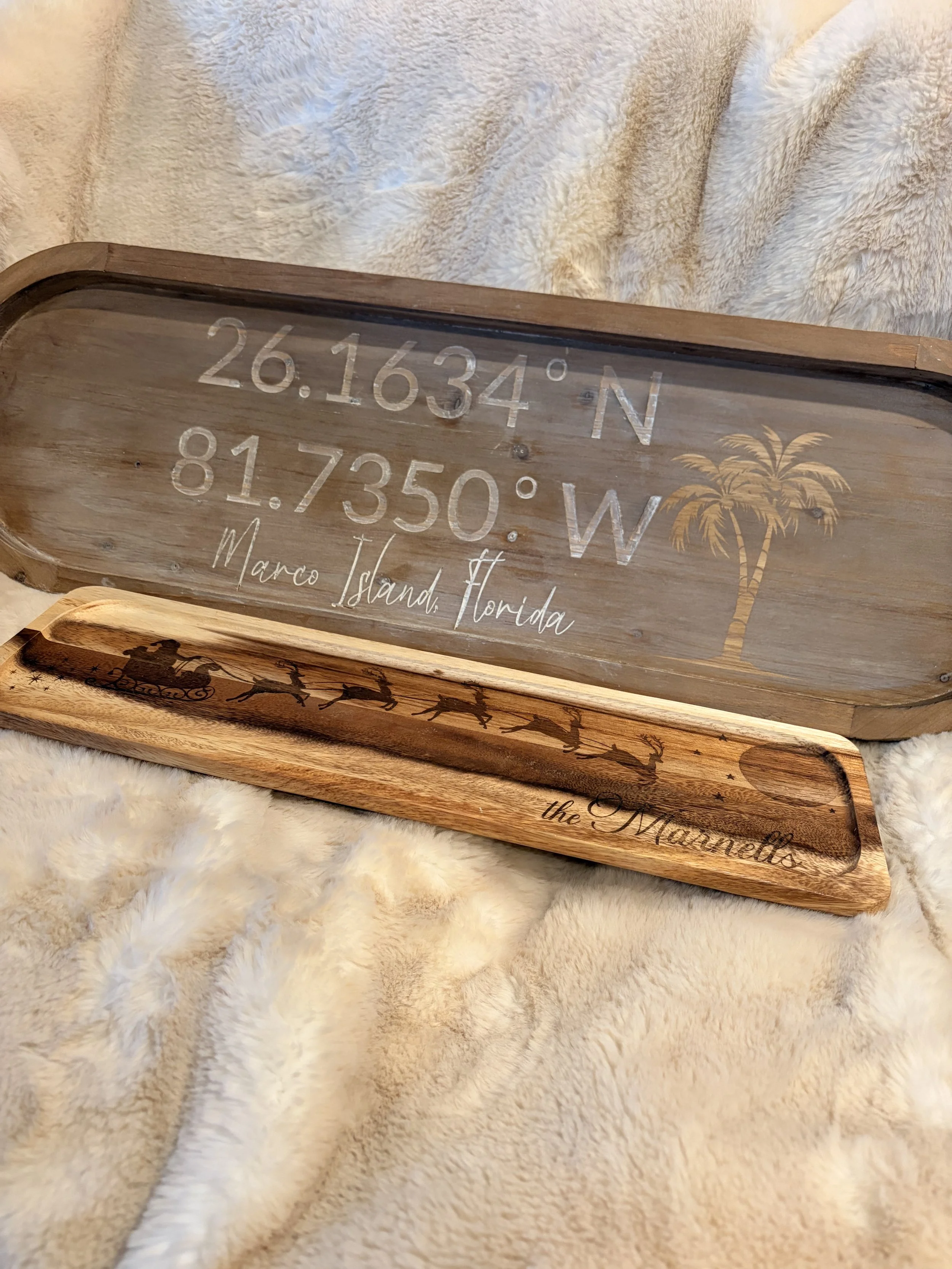 Wooden decorative tray with GPS coordinates, location name, a palm tree graphic, and a silhouette of Santa's sleigh with reindeer, on plush beige fabric.