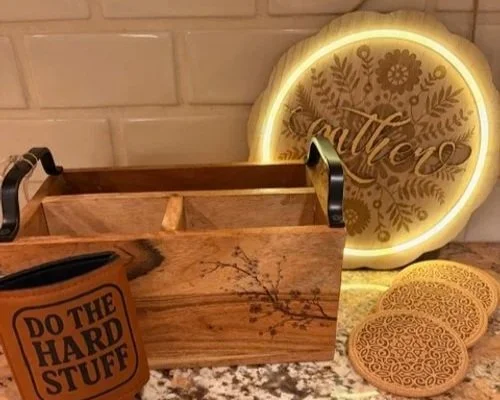 A wooden box with black handles, a lit round sign with floral and scripting design reading "Mother," three decorative cookies with intricate patterns, and a container with a label that says "Do the Hard Stuff" inside a room with a brick wall.