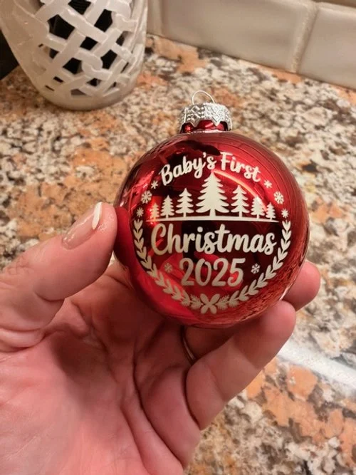 Red Christmas ornament with white lettering and design, reading 'Baby & Furst Christmas 2025,' held in a person's hand over a granite countertop.