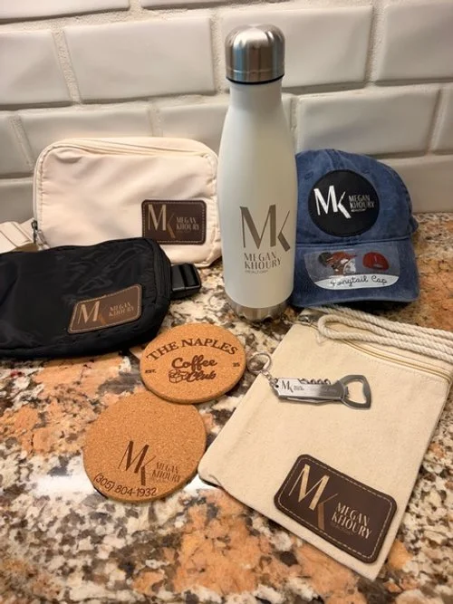 Table with various items including a white water bottle, a blue cap with a logo, two small cork coasters, two pouches, and a cork keychain, all branded Meggan Kioury.