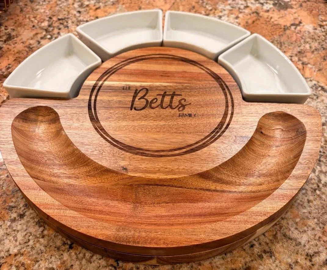 A circular wooden serving tray with a handle-like design, featuring a central area engraved with 'The Betts Family.' The tray has six small ceramic dishes arranged in a semi-circle along the top edge.