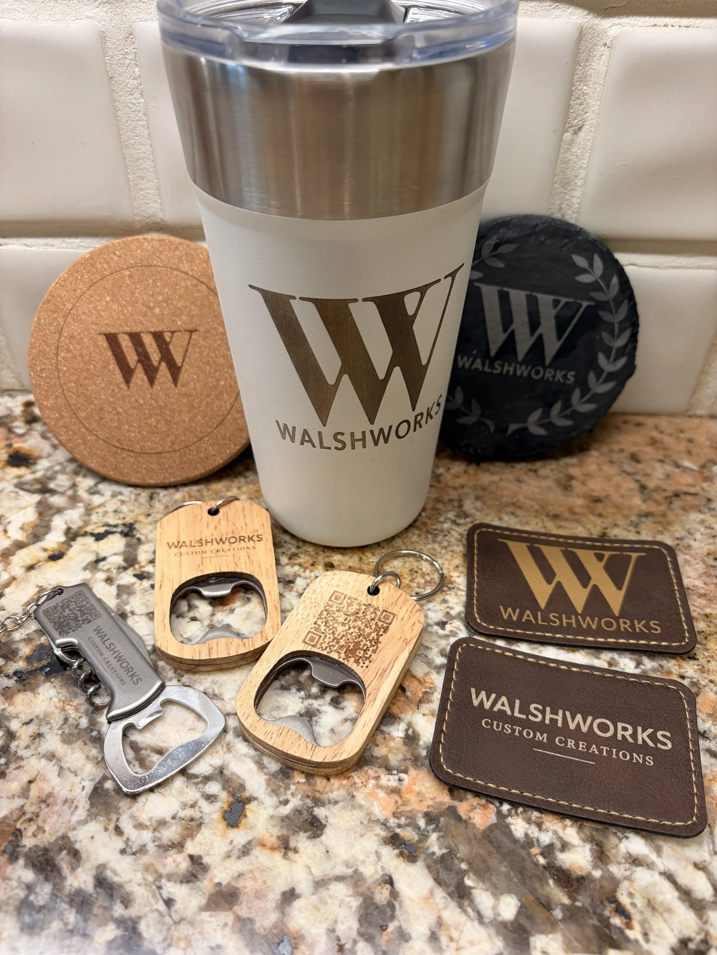 A tumbler with the Walshoworks logo, two wooden and two metal bottle openers with Walshoworks branding, two leather RFID cardholders with Walshoworks branding, and a set of coasters with Walshoworks branding on a speckled granite countertop against a white tile wall.