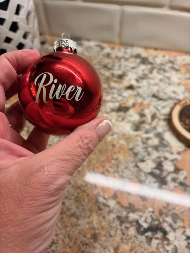 Close-up of a hand holding a shiny red Christmas ornament with the word 'River' written on it, on a granite countertop.