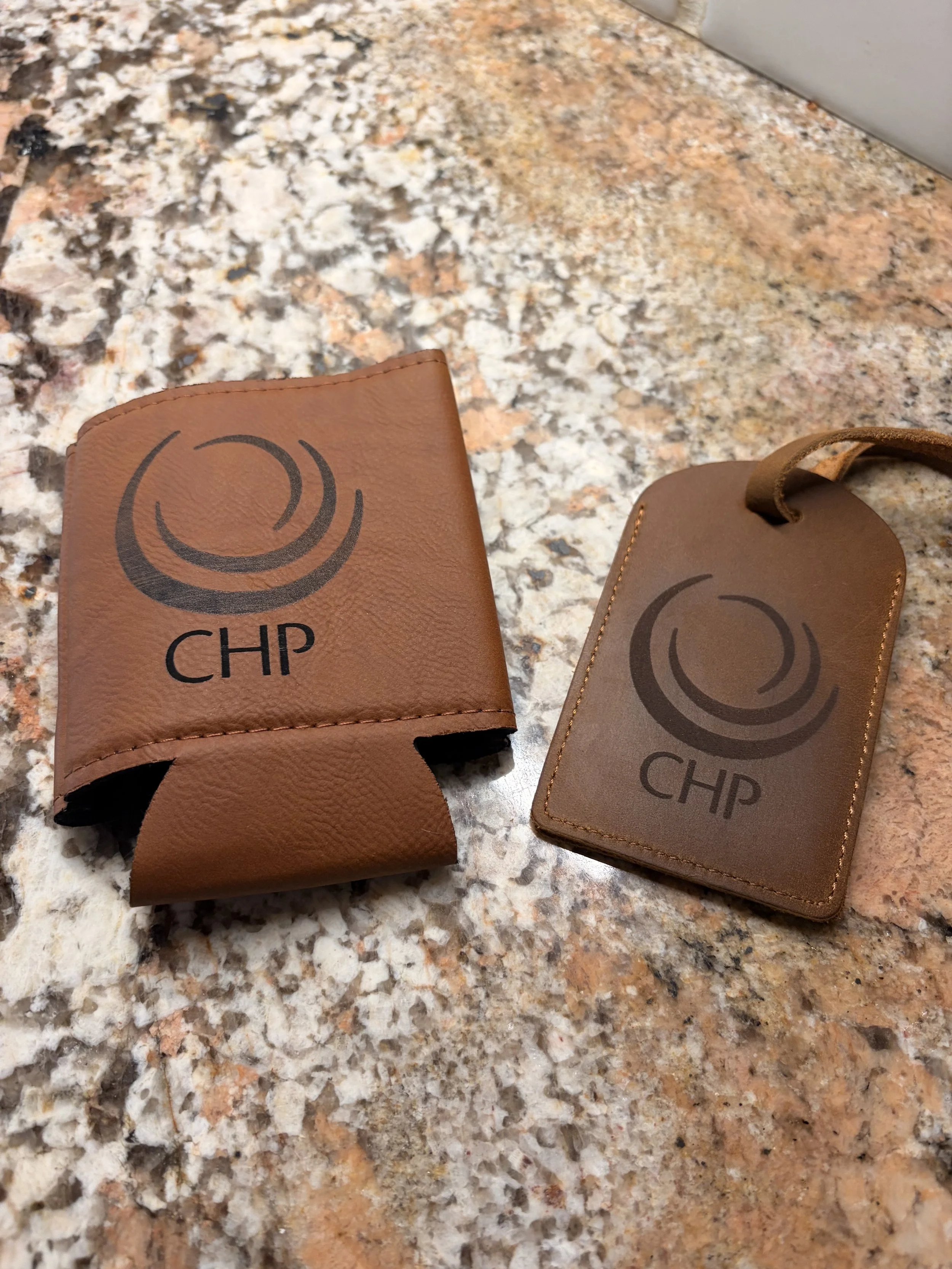Two brown leather keychain holders on a granite countertop, each with a logo and the letters 'CHP' on it.