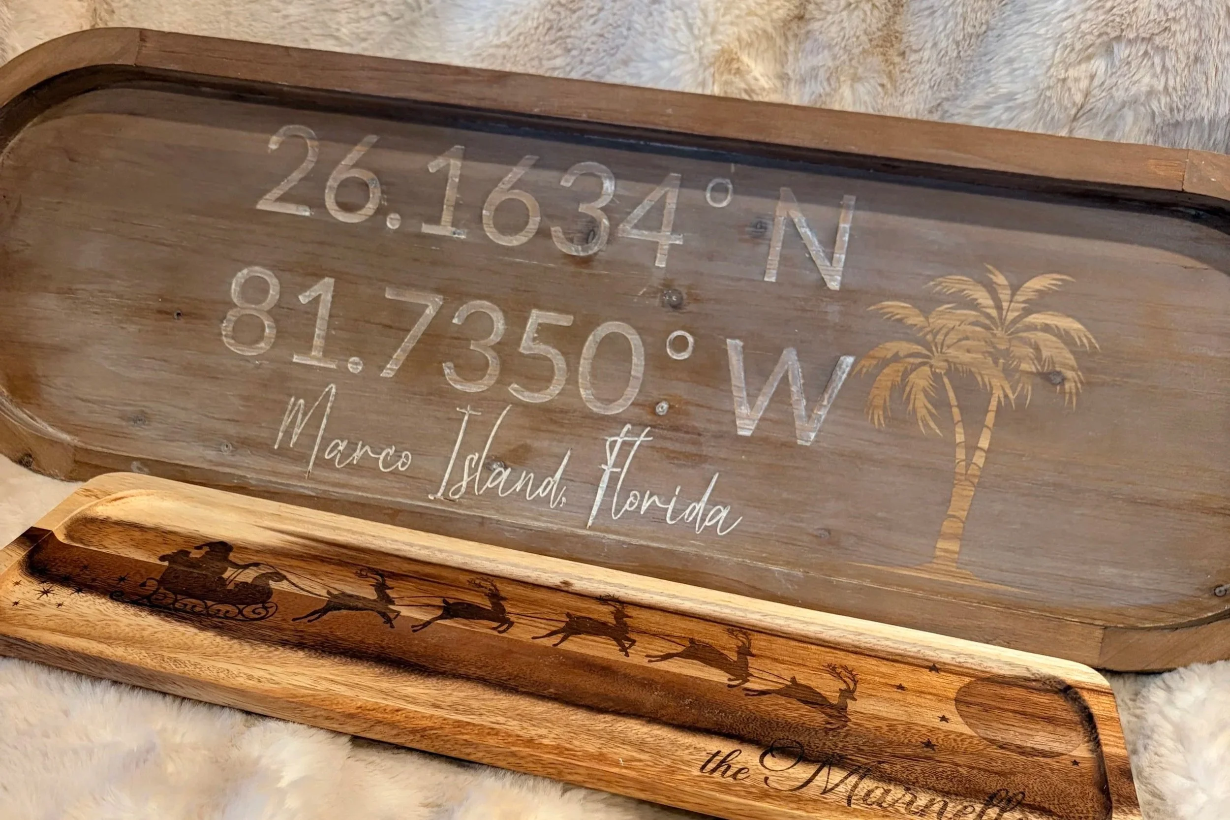 A wooden sign with engraved coordinates 26.16334°N, 81.7350°W, and the text "Marco Island, Florida" along with a palm tree illustration. A smaller wooden tray with a silhouette of Santa Claus in a sleigh pulled by reindeer is in front of the sign, placed on a soft, furry surface.