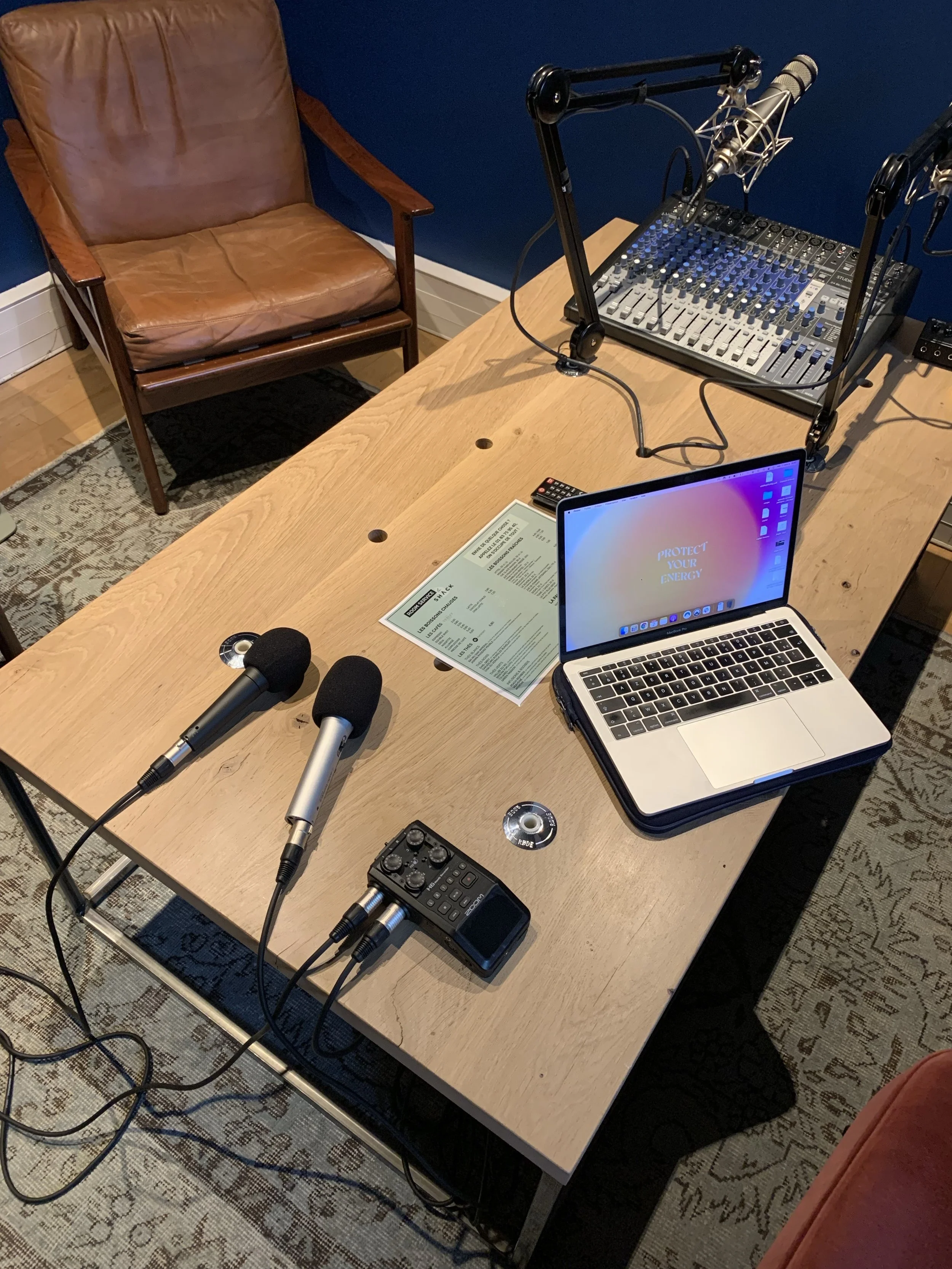 A podcast recording setup on a wooden table with two microphones, a laptop, a audio mixer, a small recording device, a remote control, and some sheets of paper. There is a brown leather chair against a dark blue wall, and an area rug on the floor.