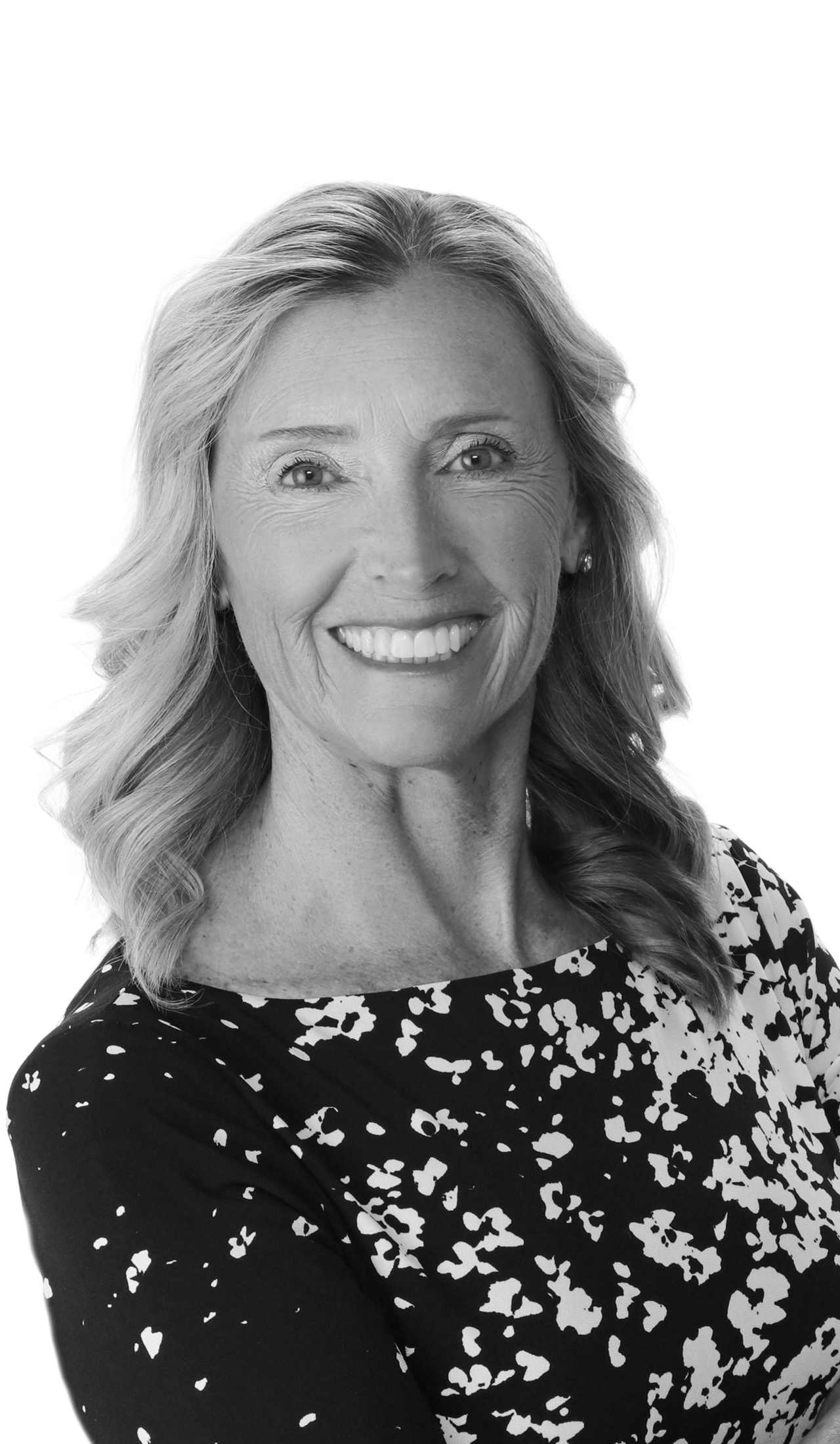 A smiling middle-aged woman with long wavy hair, wearing a black and white patterned blouse, is posing for a portrait against a plain background.