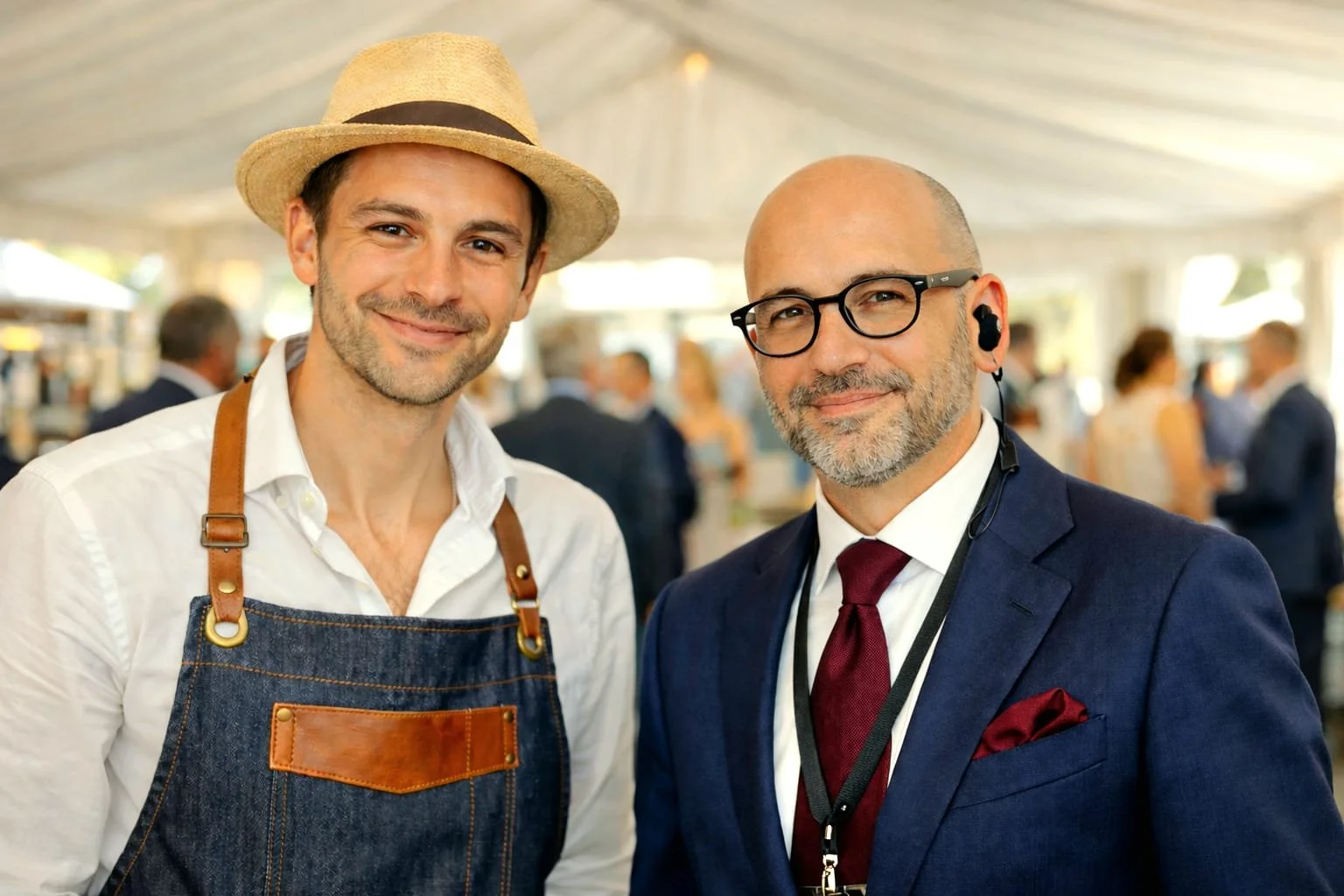 Two men dressed professionally at a social event, one wearing a white shirt, apron, and straw hat, the other in a dark suit with glasses and a burgundy tie, inside a brightly lit tent with people in the background.