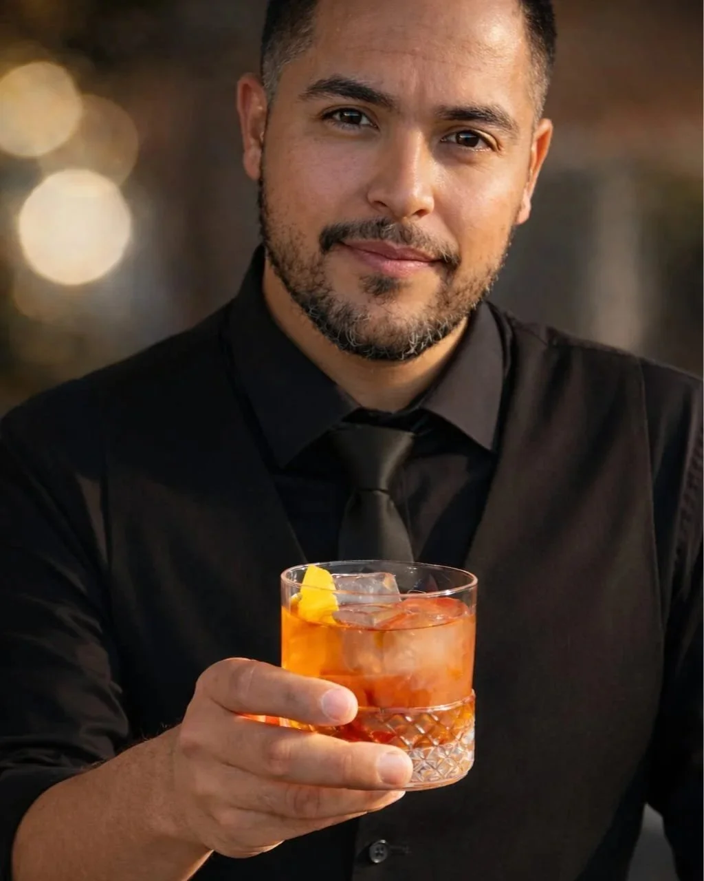 A smiling man with a beard in a black shirt and black tie holding a glass of orange cocktail with ice and an orange slice.