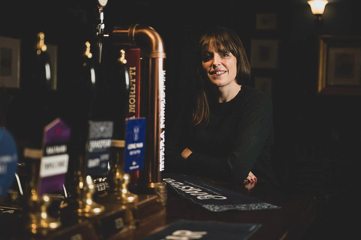 A woman with long brown hair and a black shirt sitting at a bar with beer taps in the foreground, dim interior with framed pictures on the wall and warm lighting.