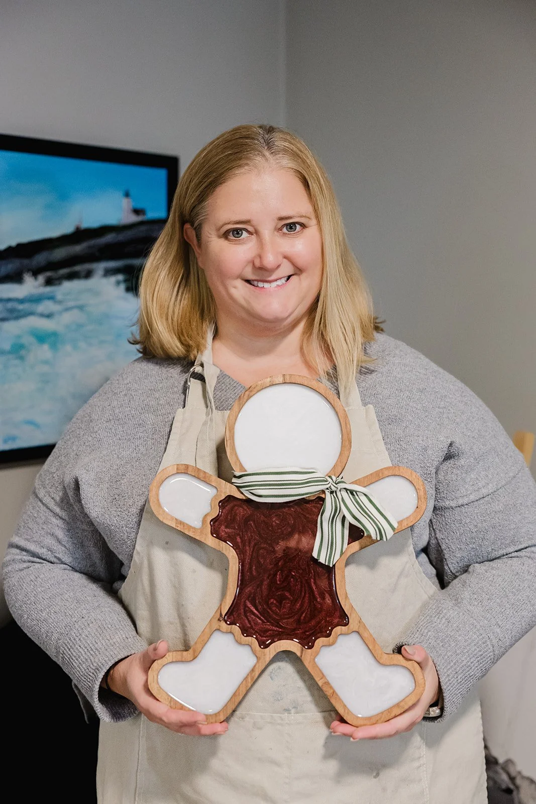 A smiling woman with blonde hair wearing a gray sweater and beige apron, holding a large gingerbread man-shaped ceramic plate with a ribbon tied around its neck, standing in a room with an ocean painting in the background.