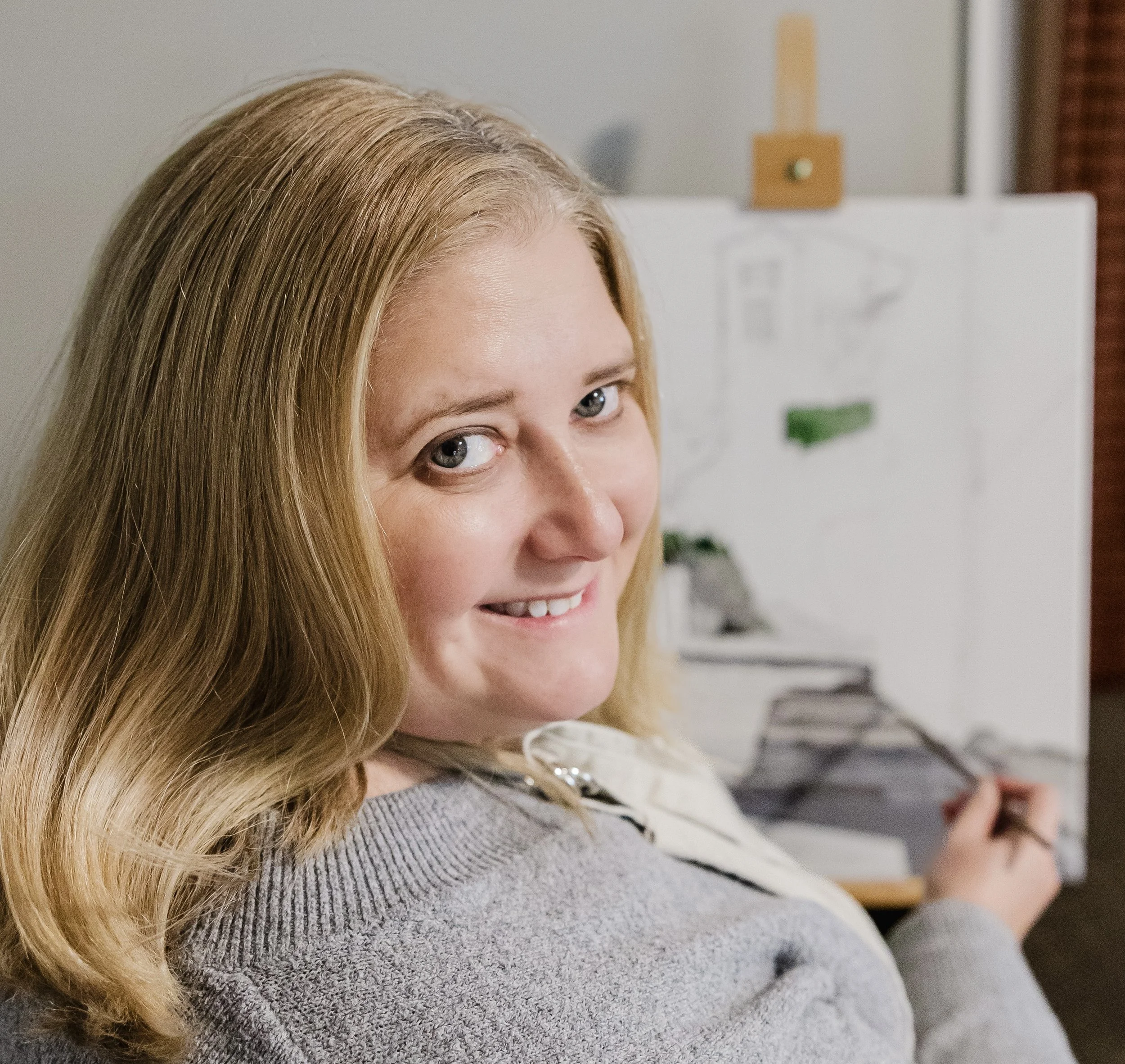 A woman with long blonde hair smiling at the camera while painting a landscape on a canvas.