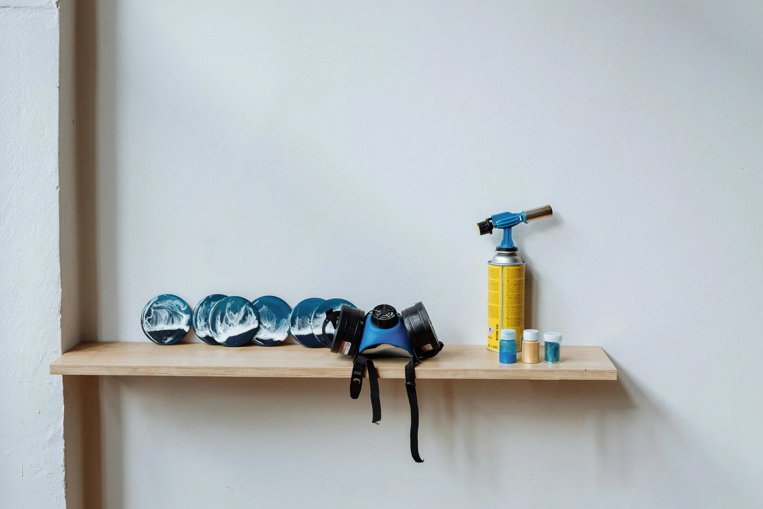 A wooden shelf on a white wall holding a gas mask, a spray paint can, and several bottles of paint or spray.
