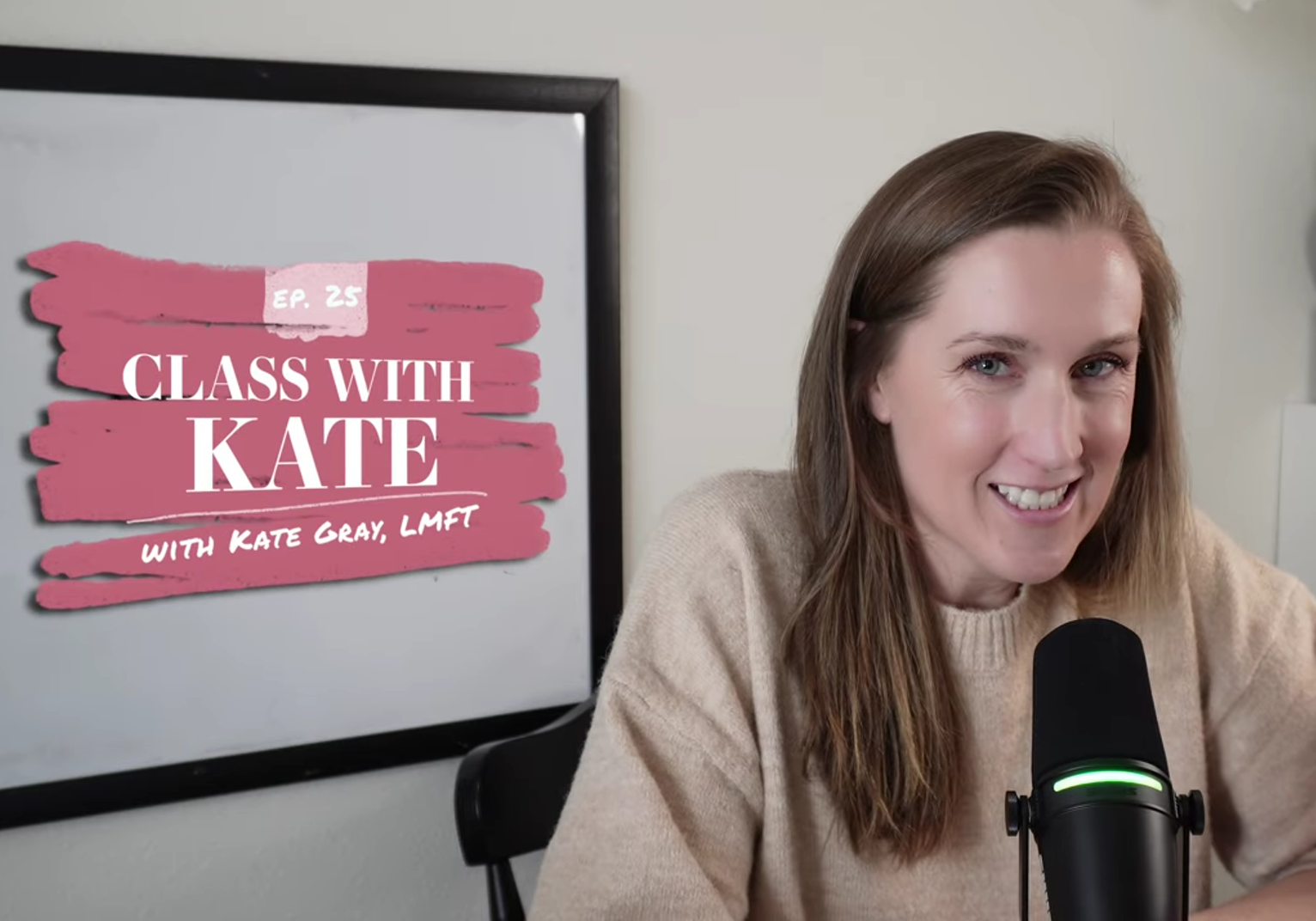 A woman with brown hair smiling at a microphone in front of her, in a room with a white wall and a black chair, with a television screen in the background displaying a graphic that says 'Episode 25, Class with Kate, with Kate Gray, LMFT'.