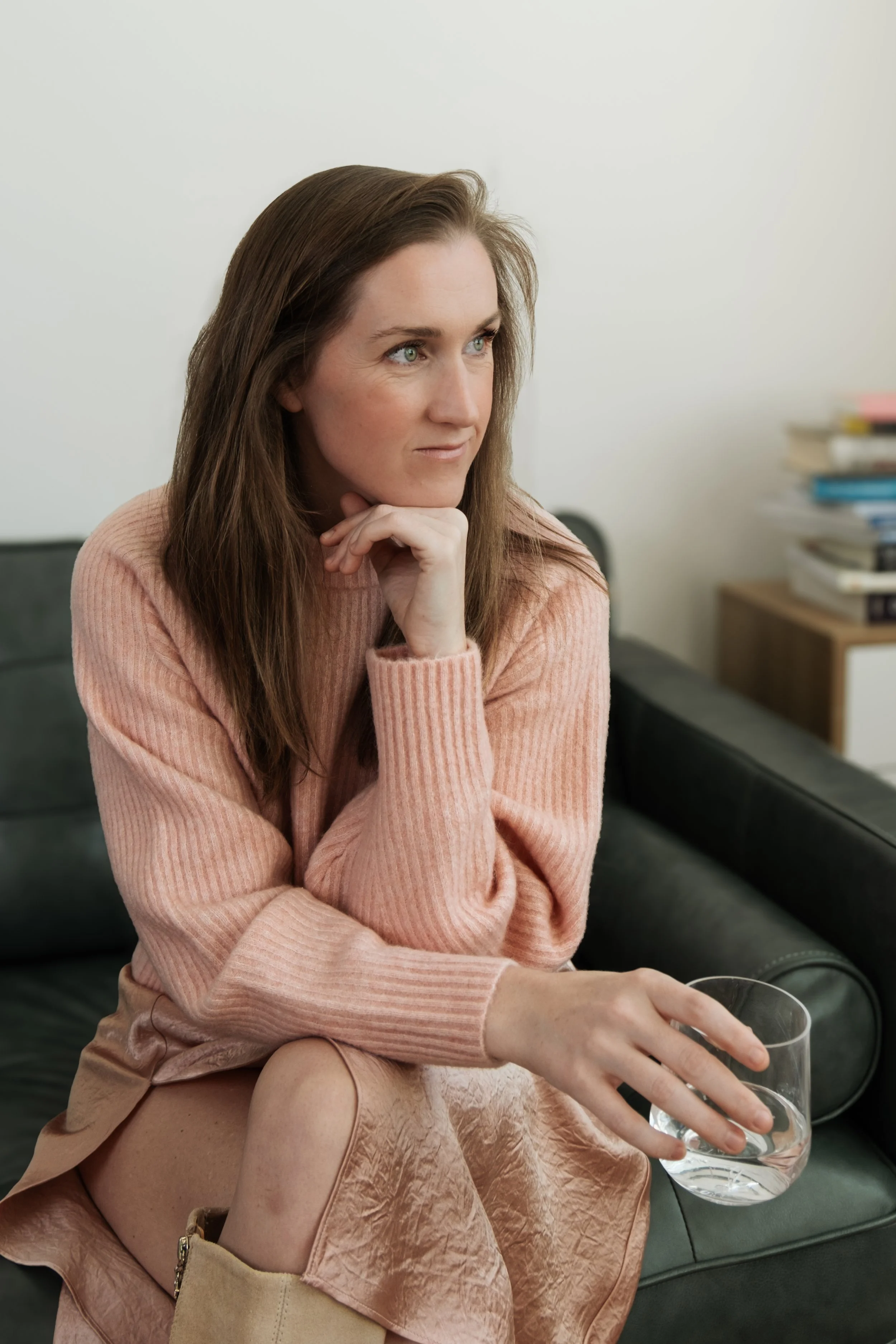A woman with long brown hair, wearing a pink sweater, sitting on a green couch in a room with a stack of books beside her, holding a glass of water.