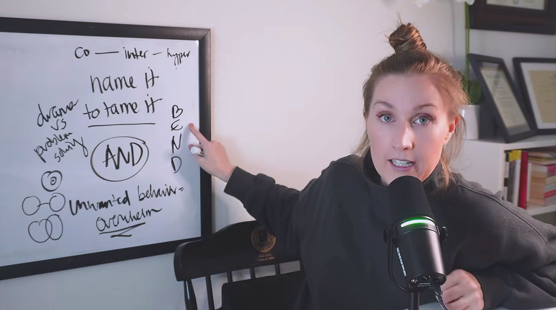 A woman with light brown hair in a bun pointing to a whiteboard with handwritten notes about ADHD and related topics, while speaking into a microphone.