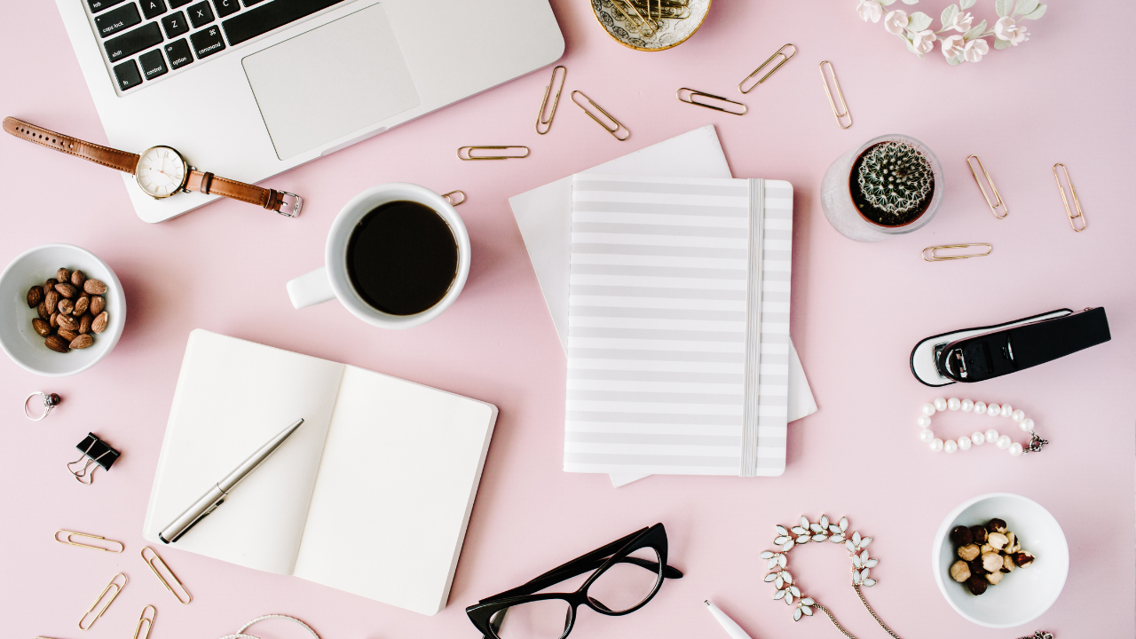 Flat lay of a pink workspace with a laptop, coffee cup, notebooks, glasses, pens, paper clips, jewelry, a cactus, and office supplies.